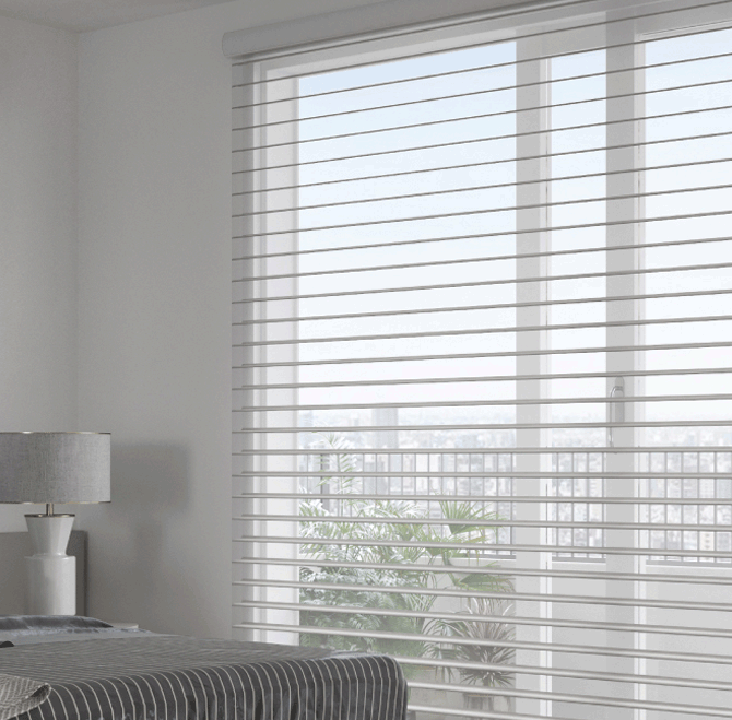 Interior view of a bedroom with sheer white blinds covering large windows, a bedside lamp, and a bed with striped bedding.