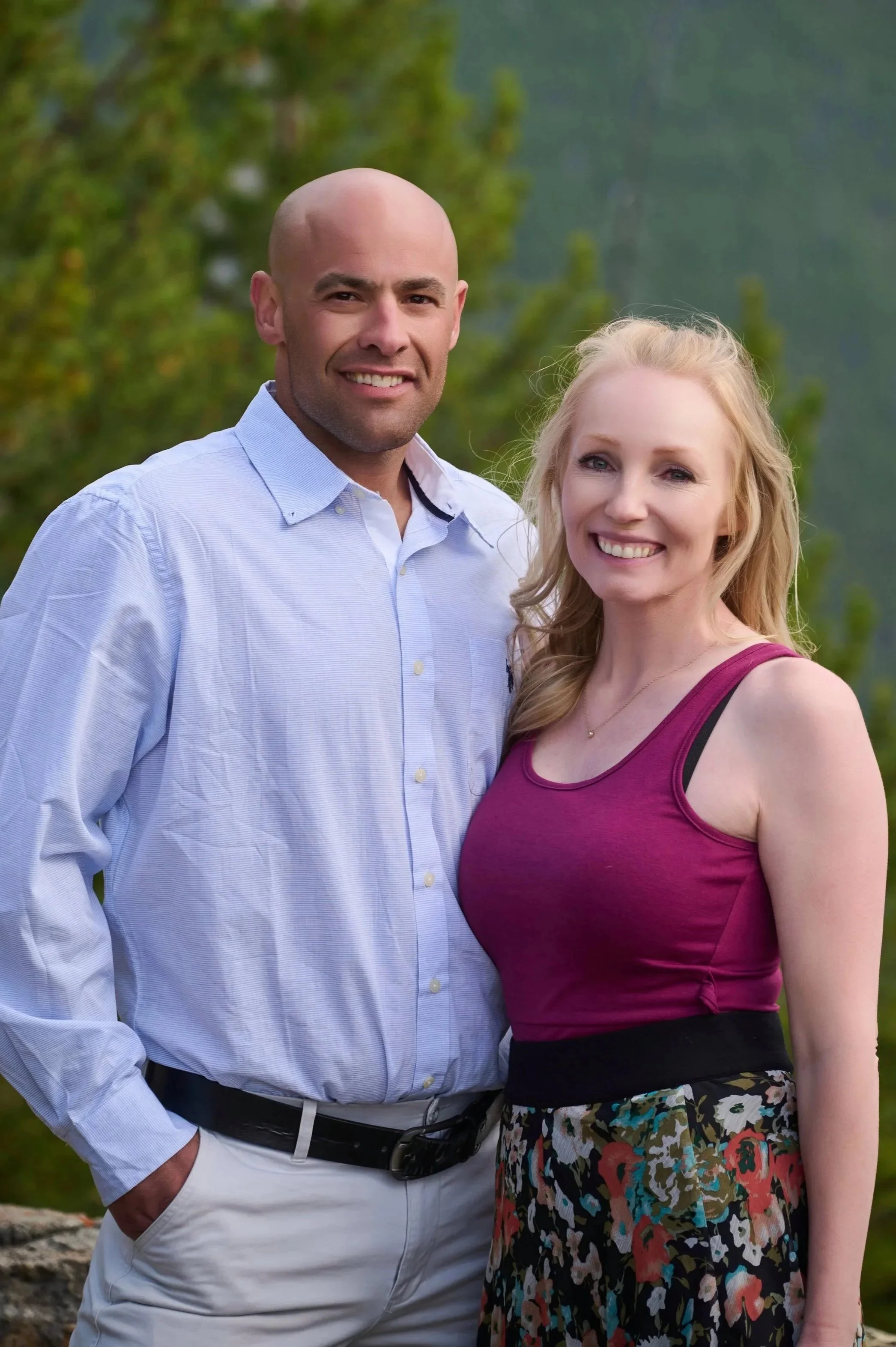 A smiling man with a bald head wearing a light blue button-up shirt and white pants, standing next to a smiling woman with long blonde hair wearing a sleeveless burgundy top and a multicolored floral skirt, outdoors with green trees in the background.