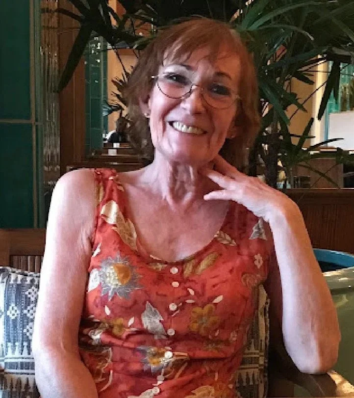 A smiling older woman with short red hair and glasses, wearing a sleeveless floral dress, sitting in a restaurant or cafe with green plants and wooden decor in the background.