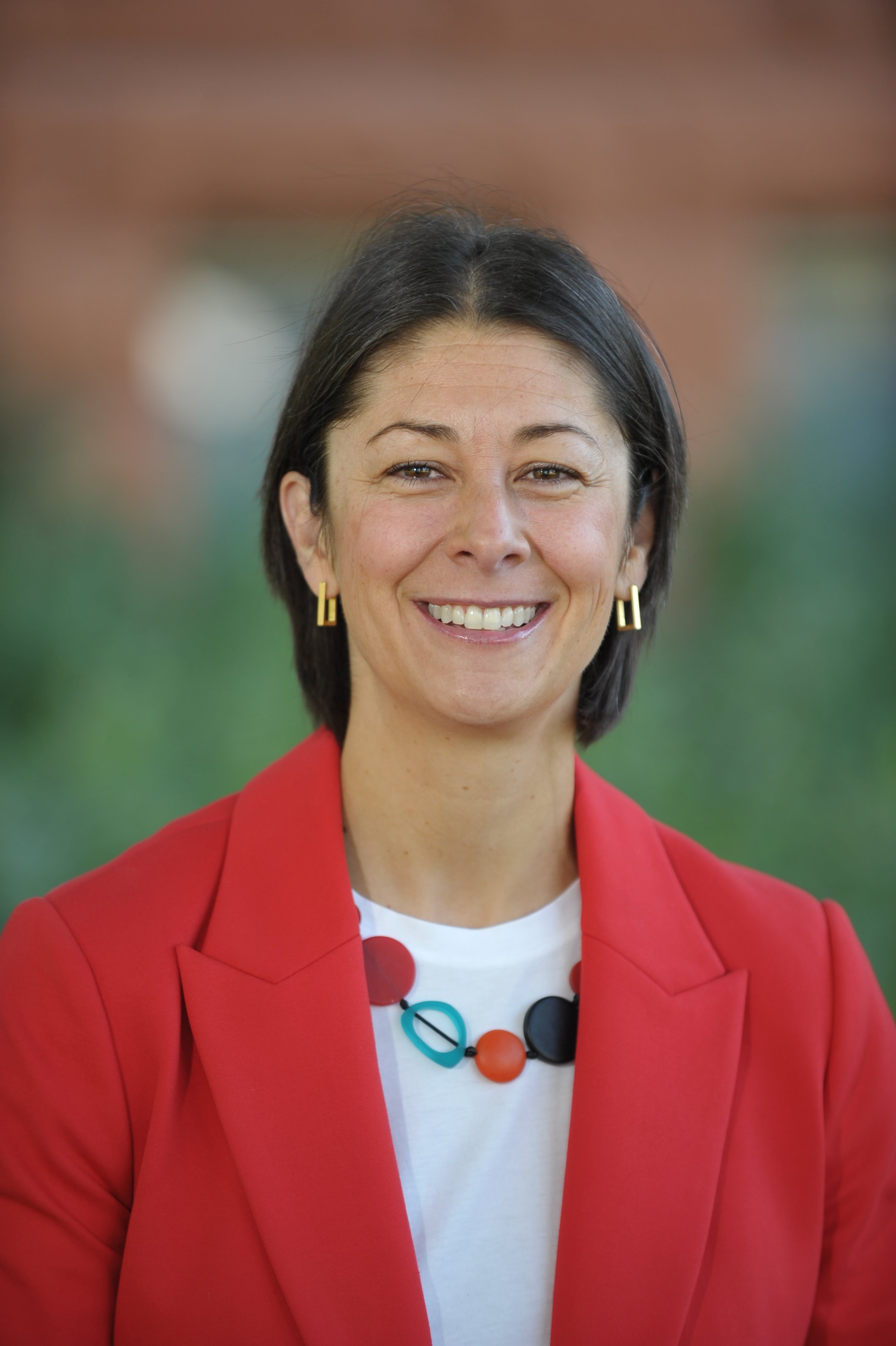 A woman with short dark hair smiling, wearing a red blazer, a white shirt with colorful polka dots, and a multicolored necklace, standing outdoors with a blurred background.