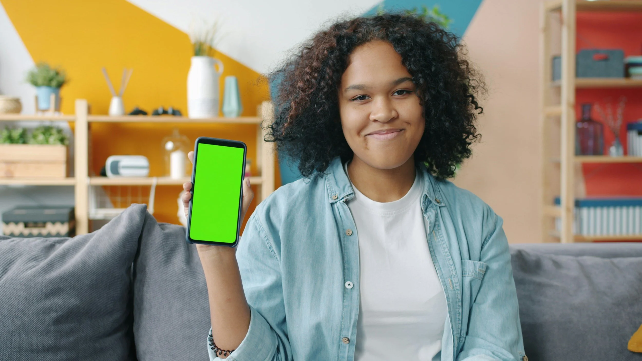 A young woman with curly hair smiling and holding a smartphone with a green screen, sitting on a gray couch in a colorful room.