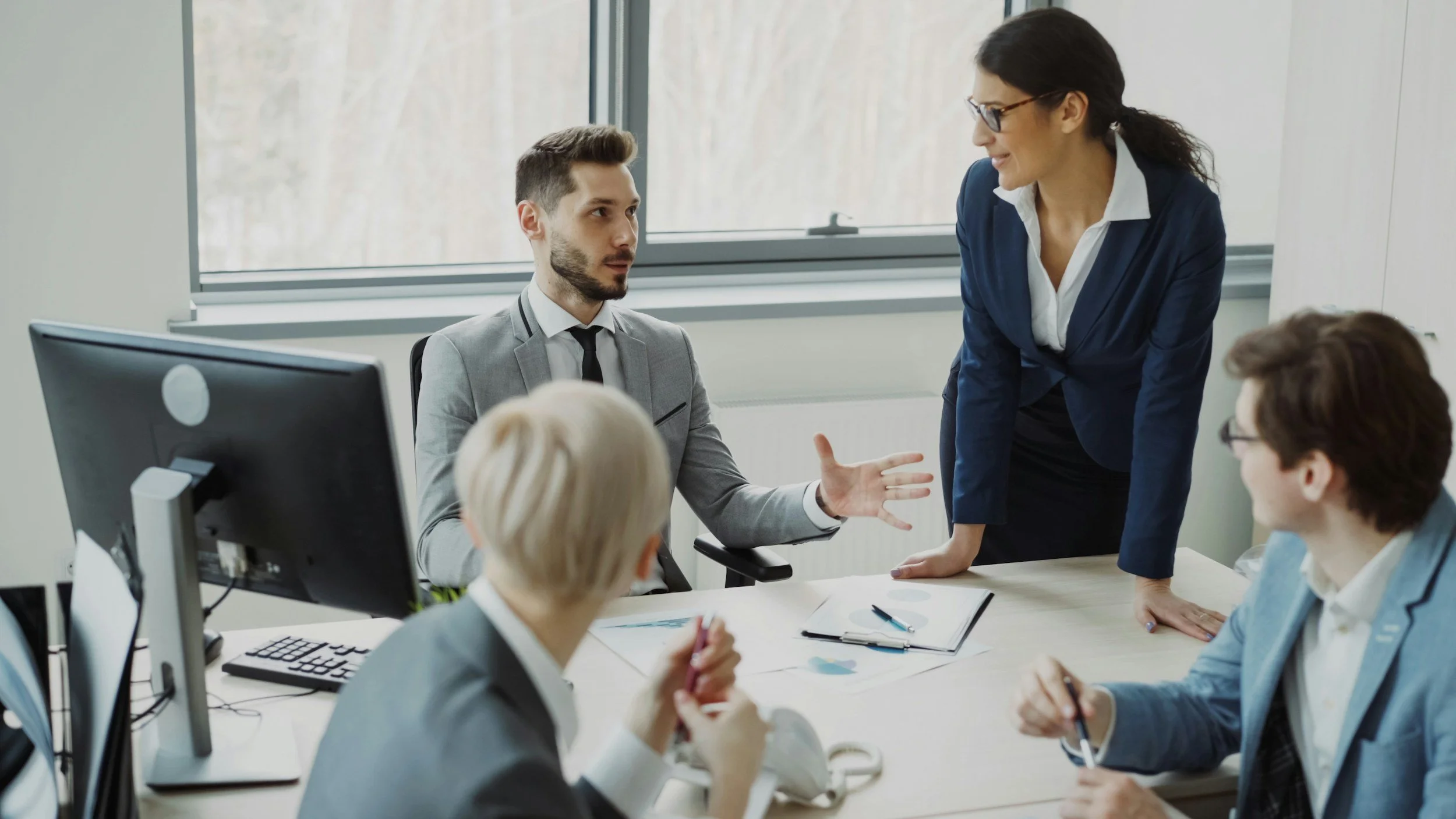 Business meeting in a conference room with five professionals, one woman standing and speaking to her colleagues at the table.