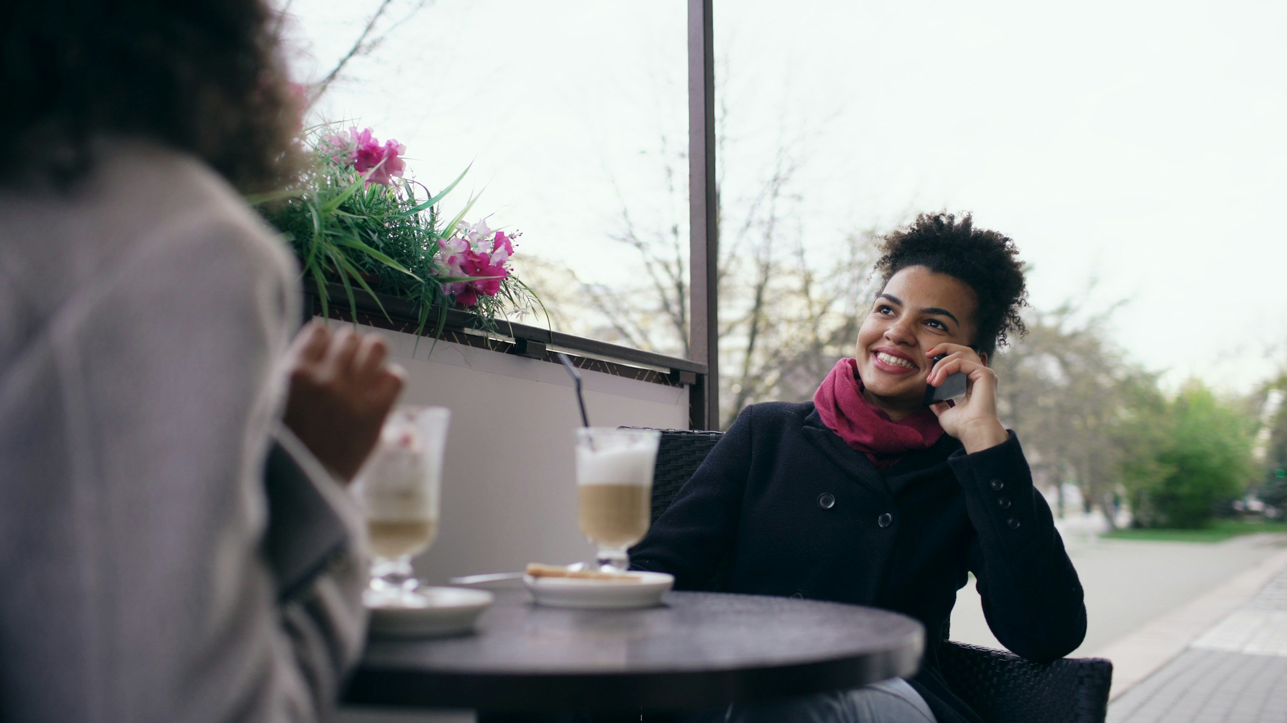 Two women sitting at a cafe table with beverages, one on the phone smiling, with a window and outdoor trees in background.