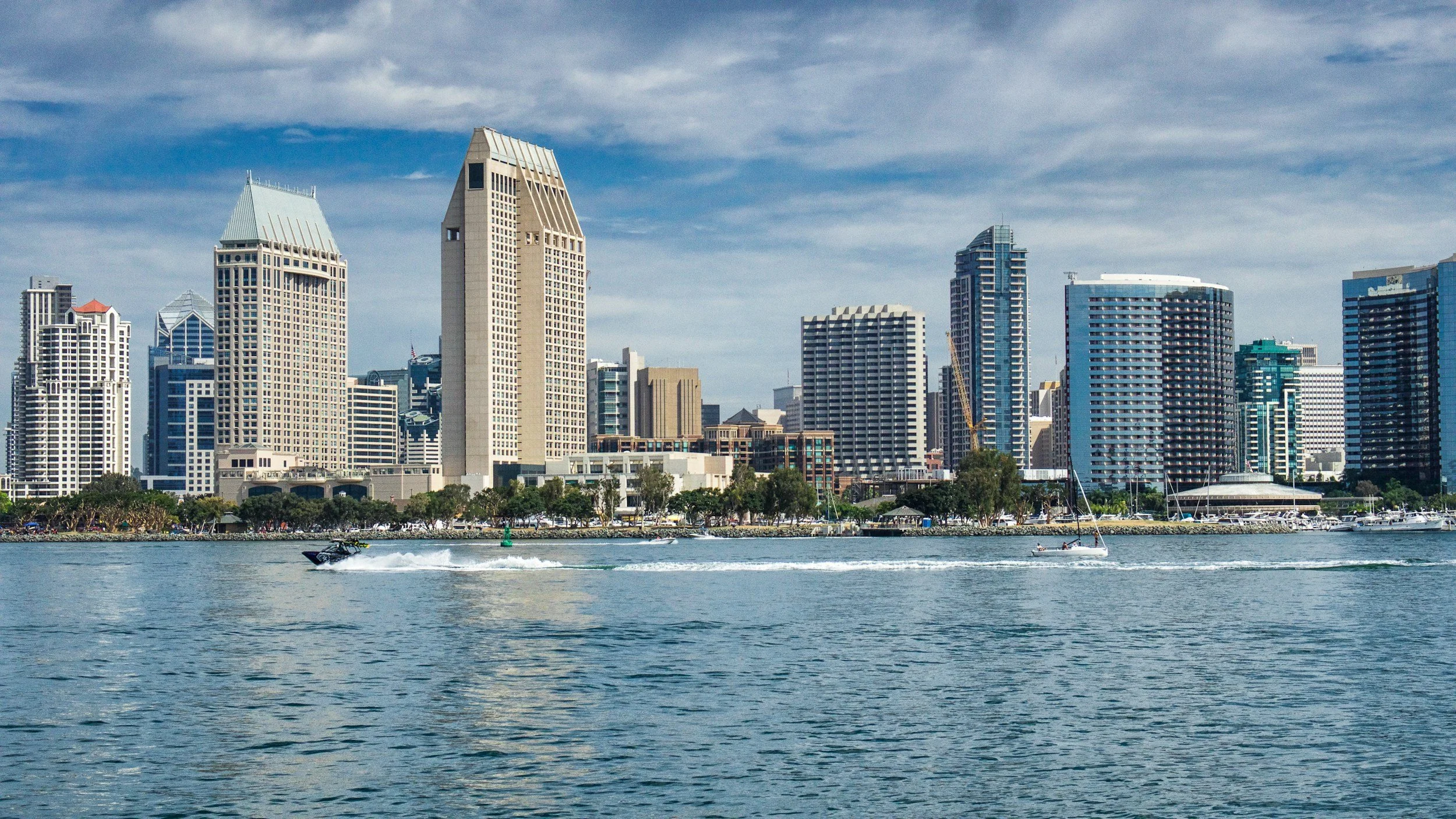 City skyline with tall buildings viewed from across a body of water with boats sailing.
