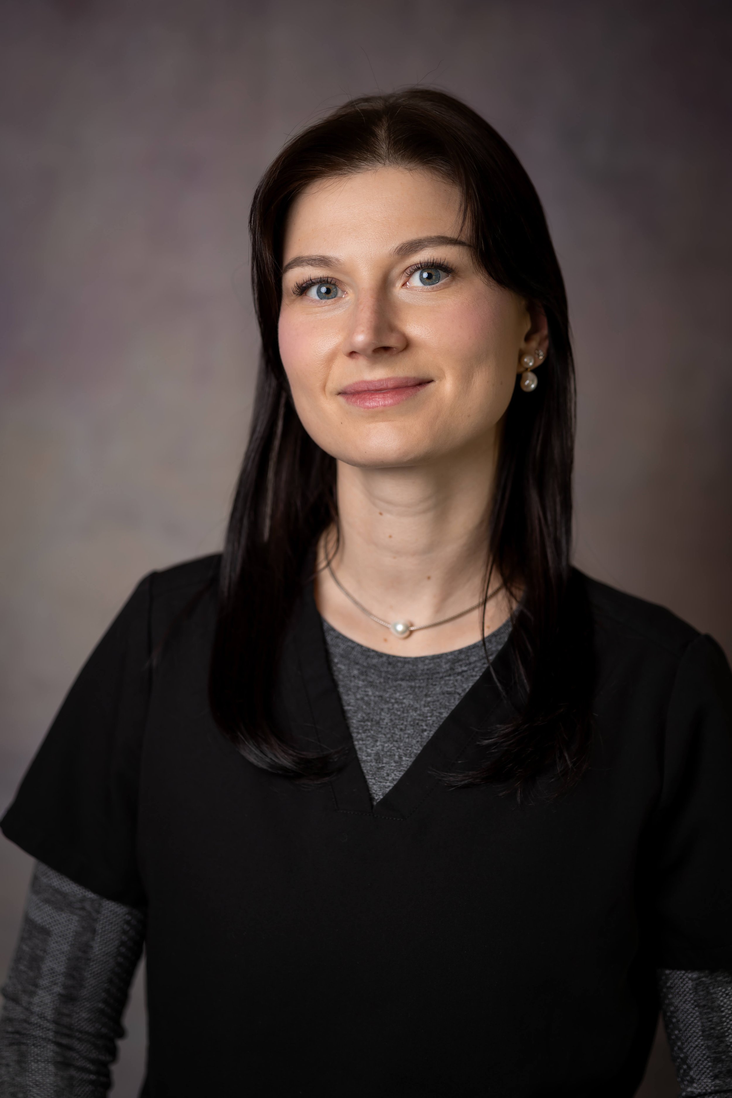 A woman with blue eyes and dark brown hair styled with a side part, wearing a black blazer over a gray top, pearl earrings, and a pearl necklace, smiling softly against a gray background.