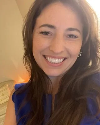 A smiling woman with long brown hair, wearing a blue top, appears to be indoors with a beige wall and warm lighting in the background.
