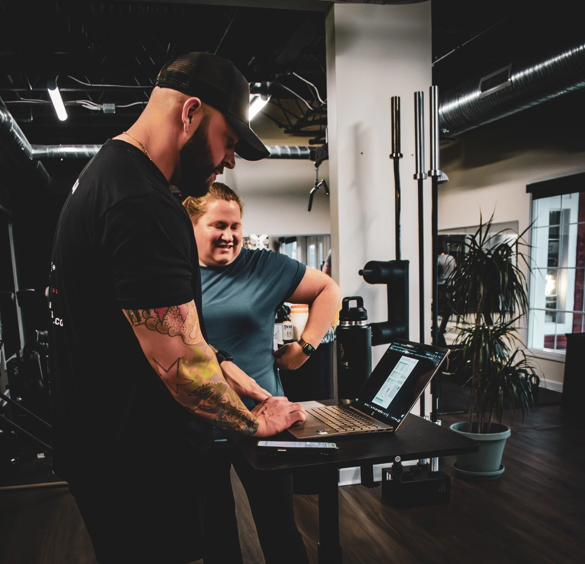 Two men working on a laptop in a gym or fitness studio, with workout equipment and a large window in the background.