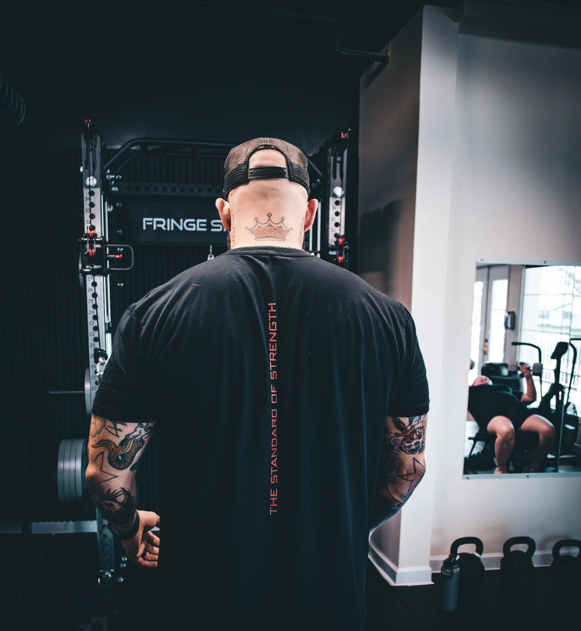 Person with tattoos on arms and tattooed neck, wearing a black shirt and a black cap backwards, standing in a gym near fitness equipment.