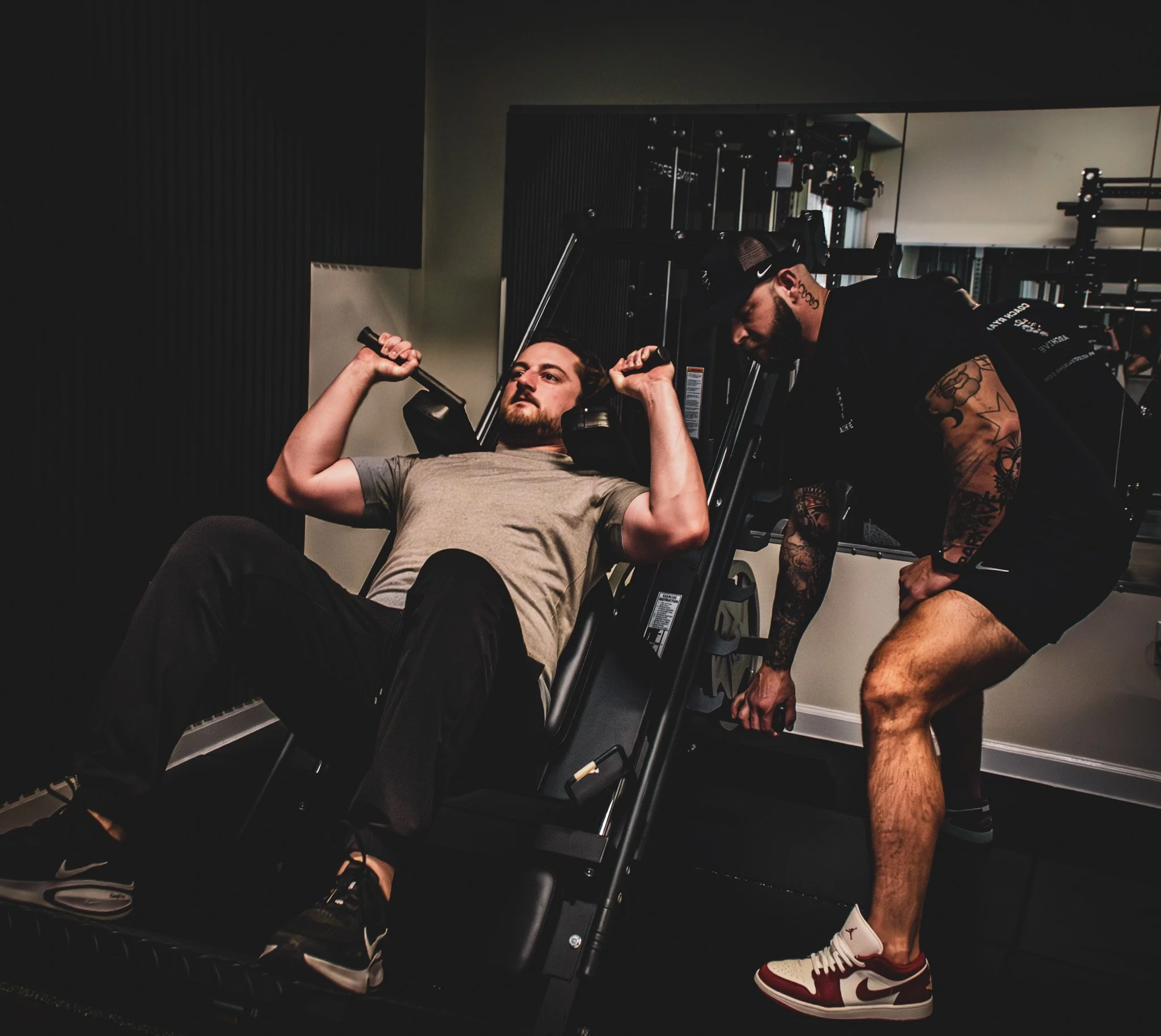 A man in a gray t-shirt and black pants lying on an exercise machine doing a shoulder press, with a trainer nearby assisting.
