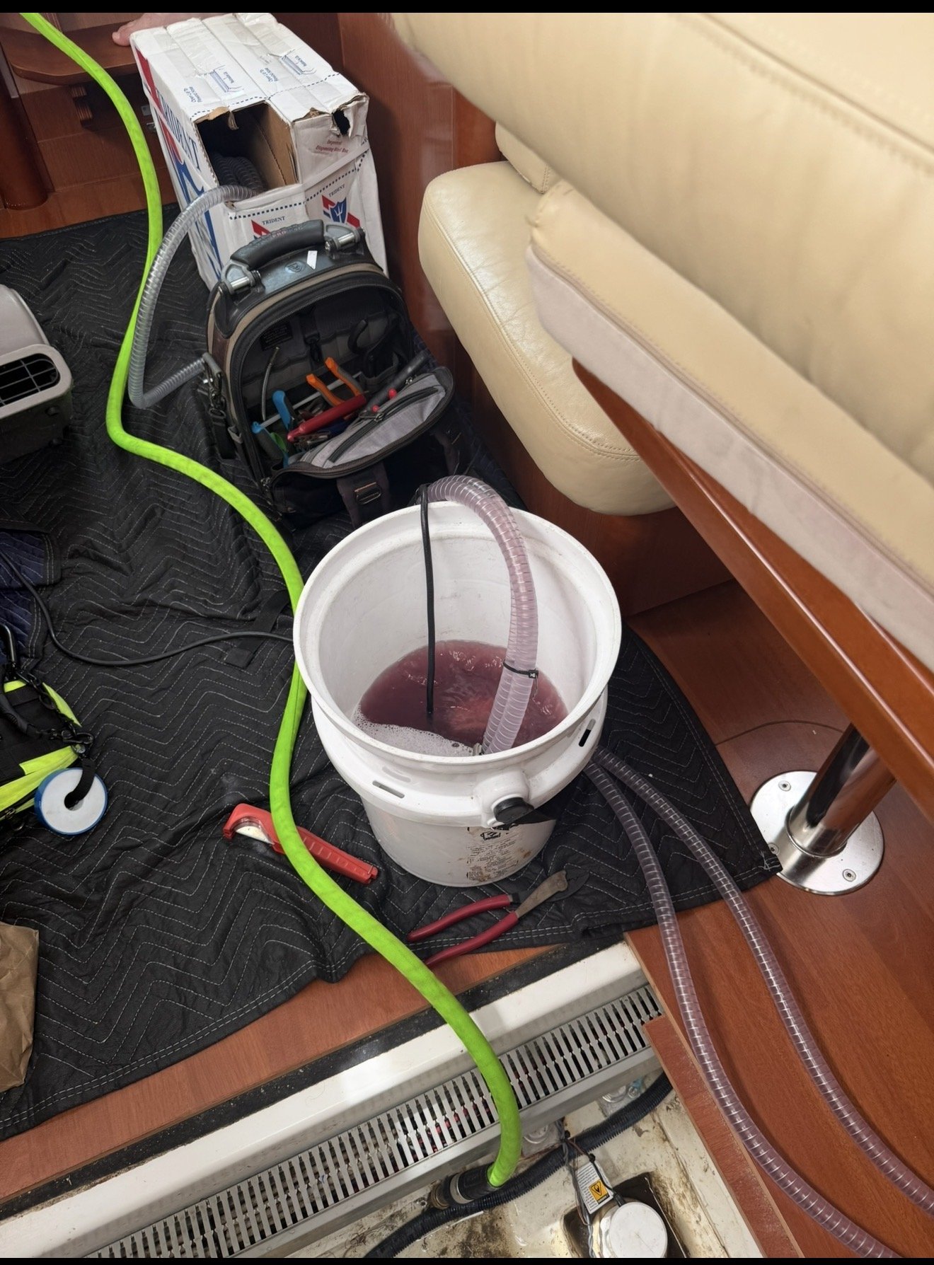 Suction equipment, hoses, tools, and a bucket of red liquid on a black pad under a wooden desk with a cream-colored seat nearby.