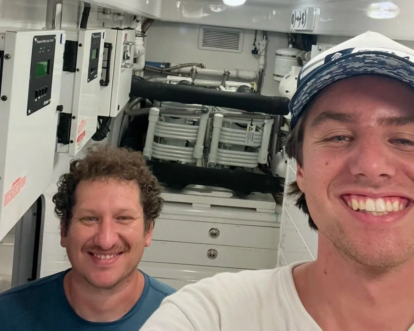 Two smiling men taking a selfie inside a technical or industrial environment with control panels and machinery in the background.