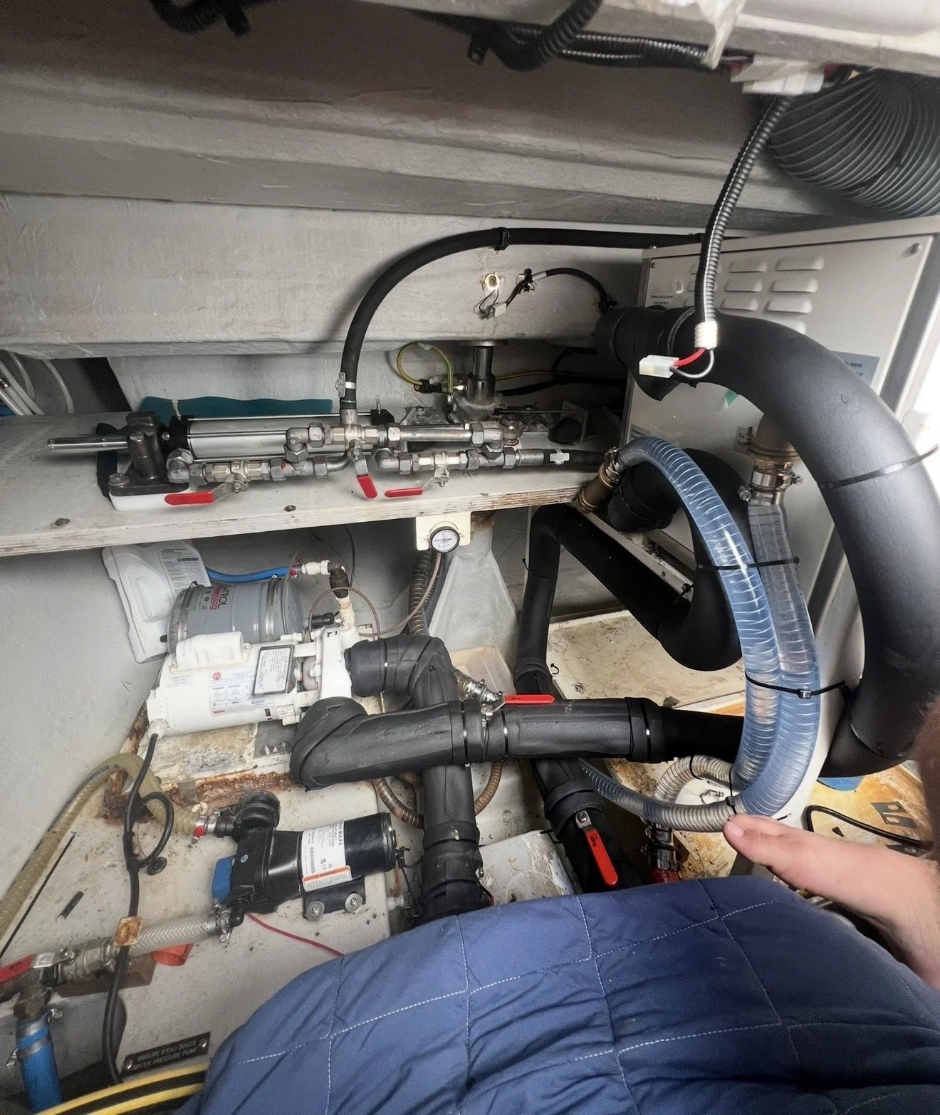 The image shows a complex setup of plumbing and equipment under a sink or appliance, including pipes, valves, and a pump, with a person's hand pointing at one of the pipes.