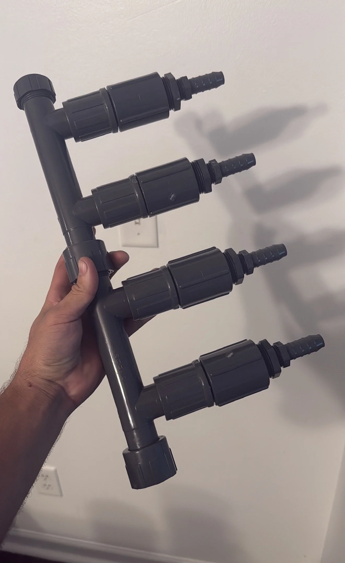 A hand holding a set of black plastic plumbing components connected together, including elbows, couplings, and a tee, against a white wall background.
