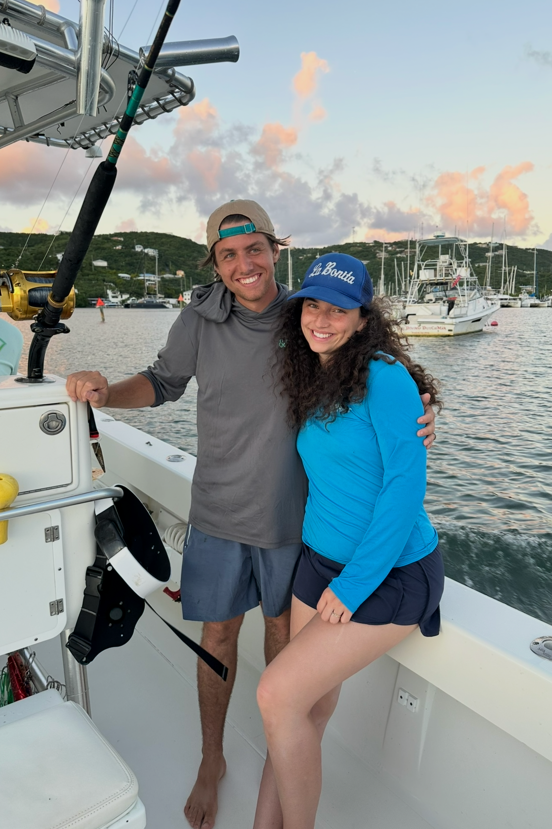 A young man and woman are smiling and posing together on a boat, with a marina and sailboats in the background during sunset.