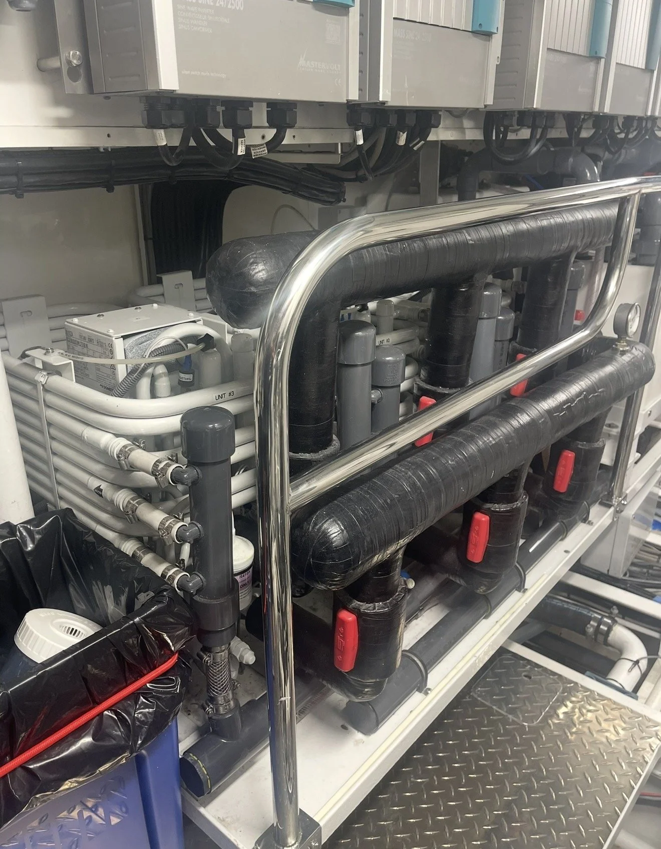 Industrial mechanical system with black pipes, white tubing, control valves, and electrical components mounted on a metal frame with a diamond plate floor.