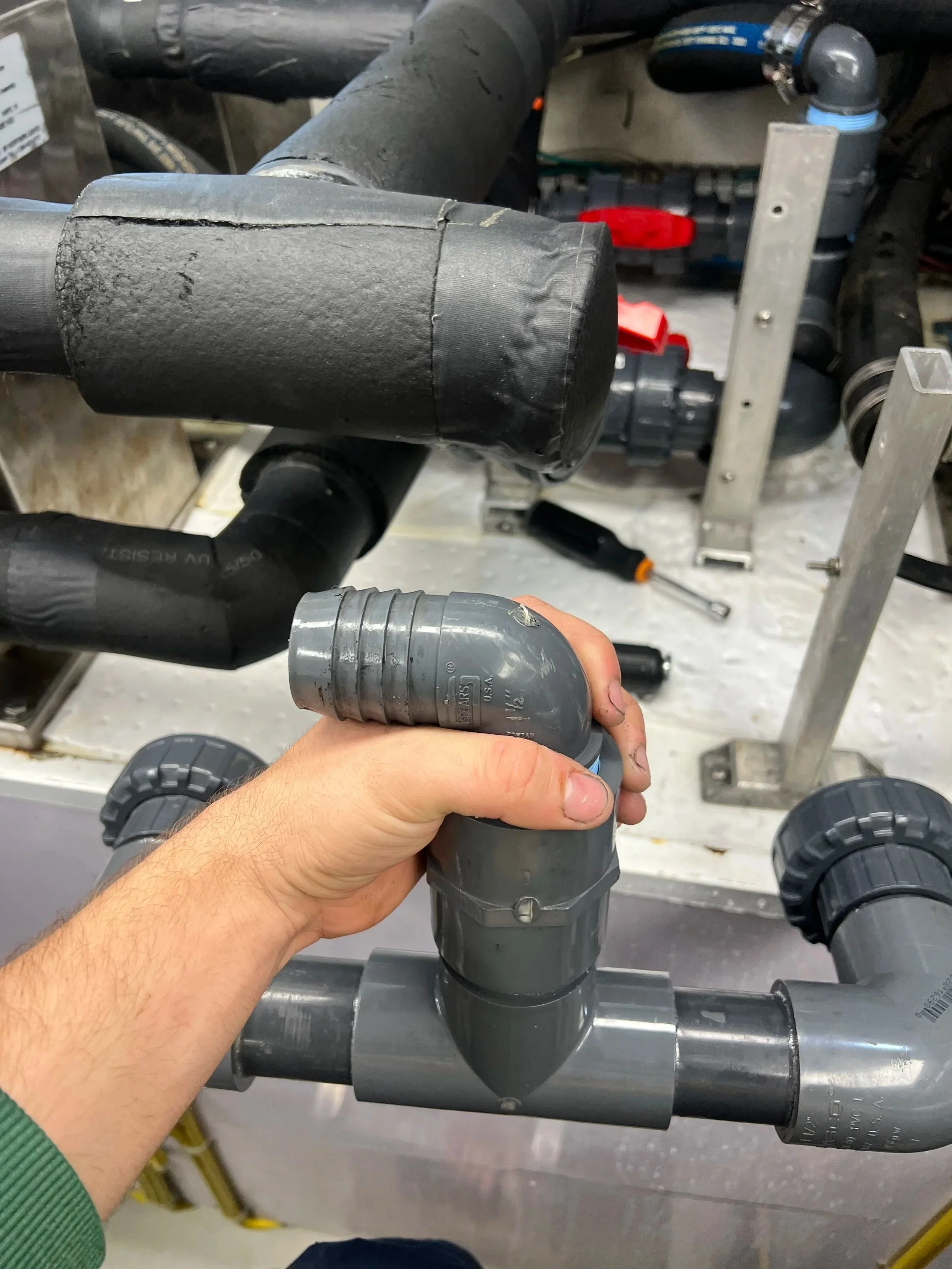 A person is holding a gray PVC pipe elbow fitting in a workshop with various pipes, tools, and equipment in the background.