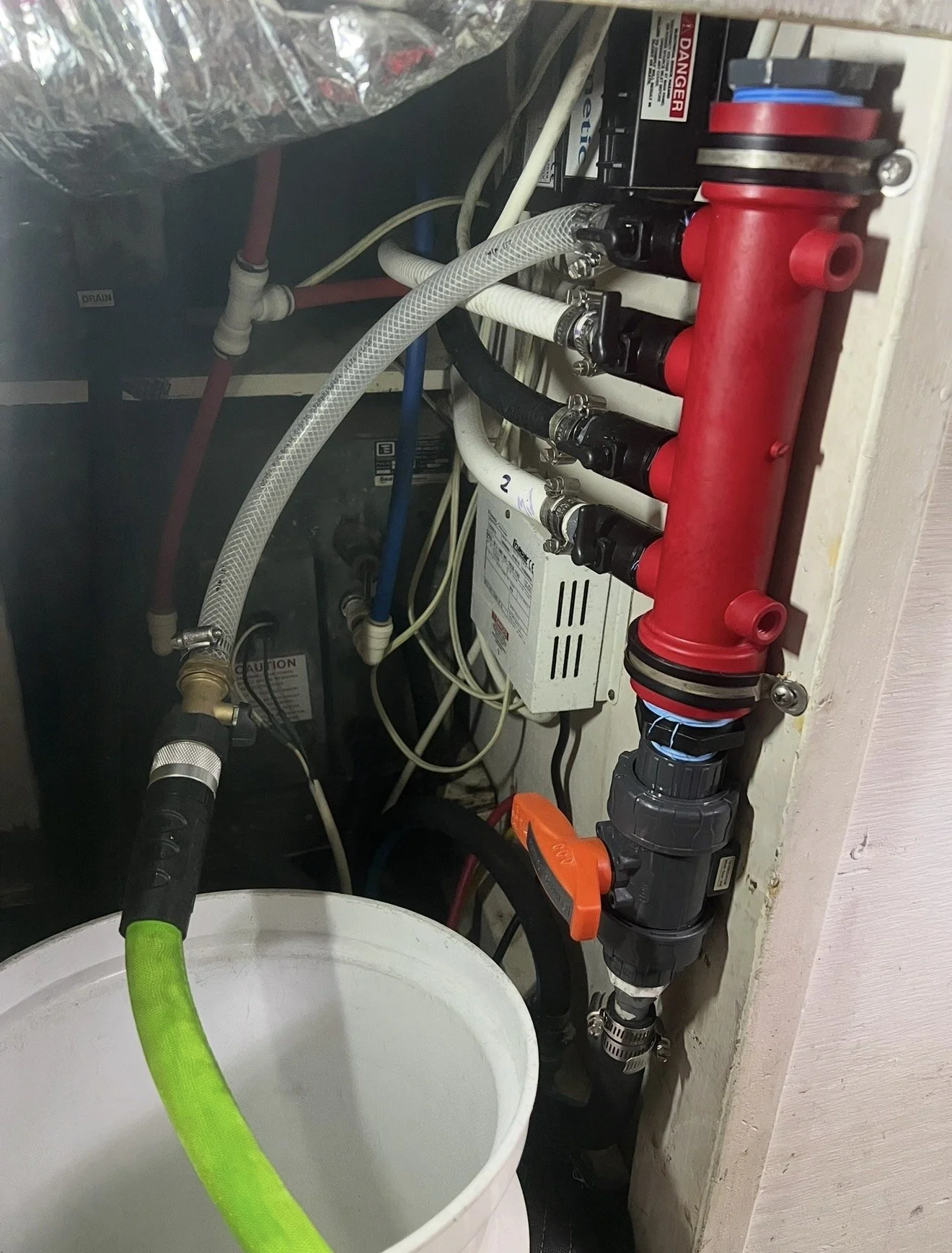 An image of plumbing inside a wall, showing a red manifold with multiple black and white hoses connected, a white box with labels, and various pipes and hoses in the background. A white bucket with a green hose is in the foreground.