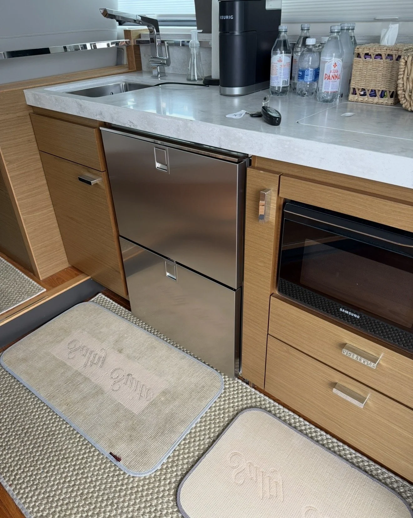 Kitchen countertop with a sink, a Keurig coffee machine, water bottles, a wicker basket, a black oven, a small stainless steel refrigerator, a key with a keychain, and beige and white floor rugs.