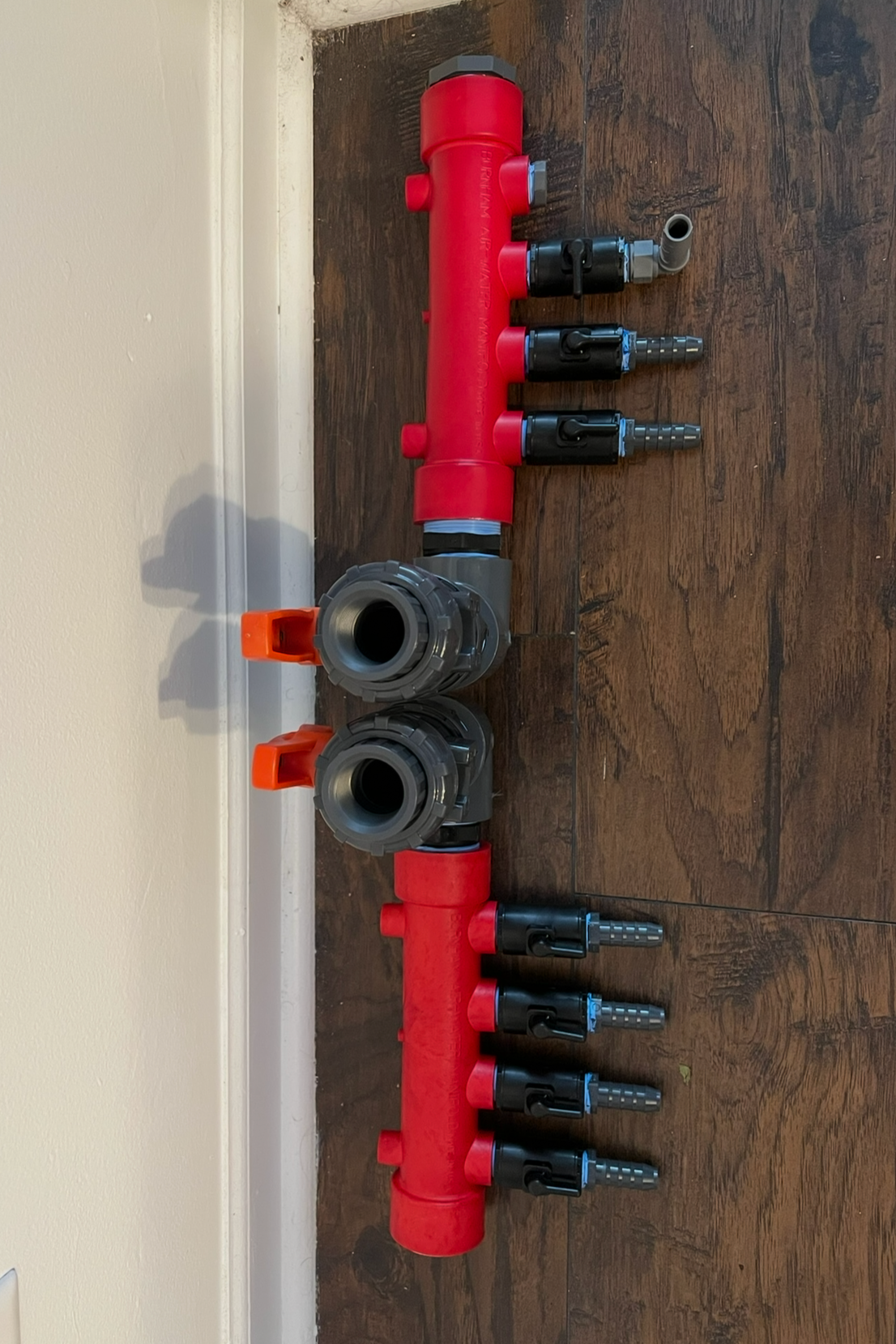 A red and gray irrigation manifold with multiple black and gray quick-connect fittings laid out on a wooden floor.