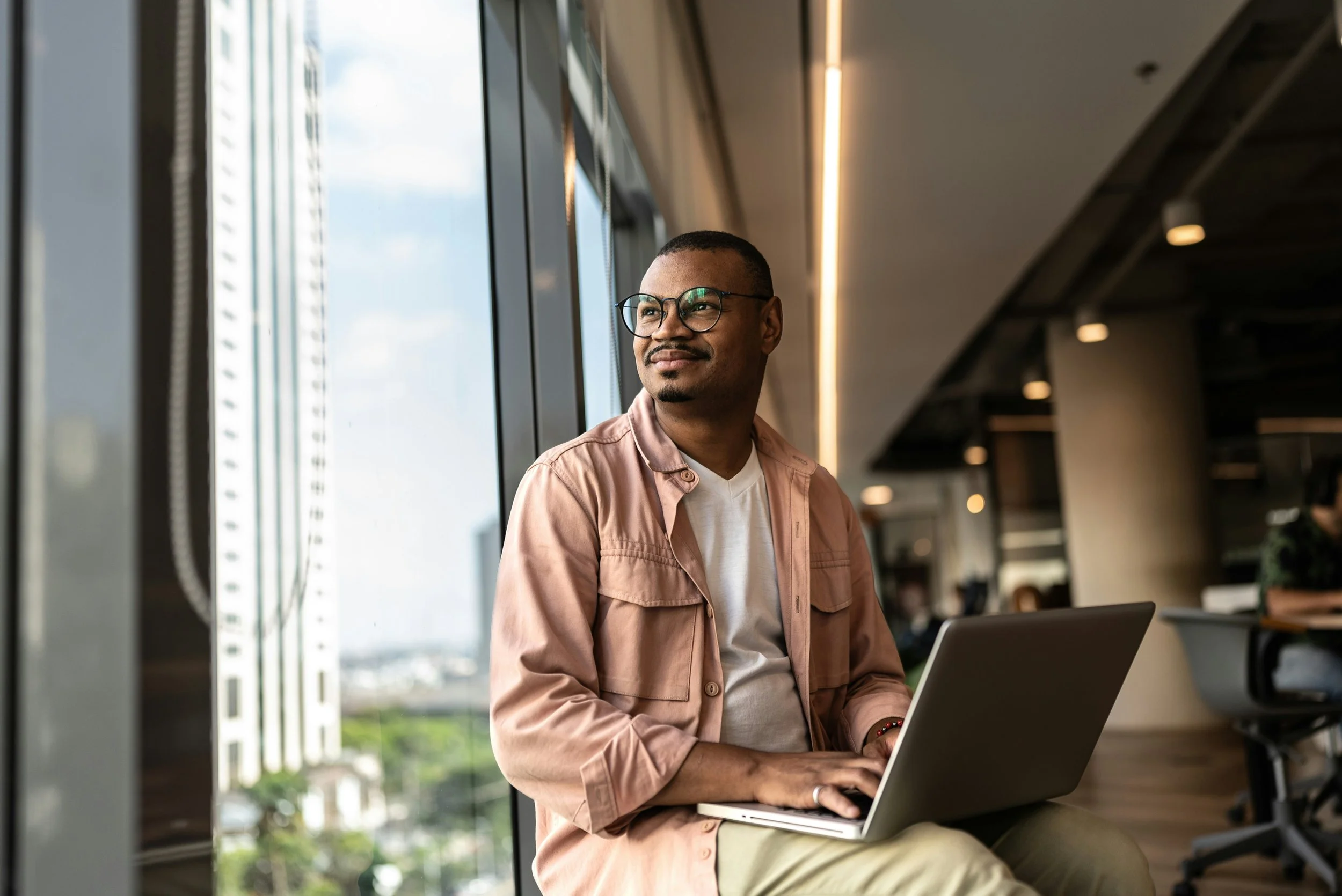A man with glasses sitting by a large window in a modern building, working on a laptop.