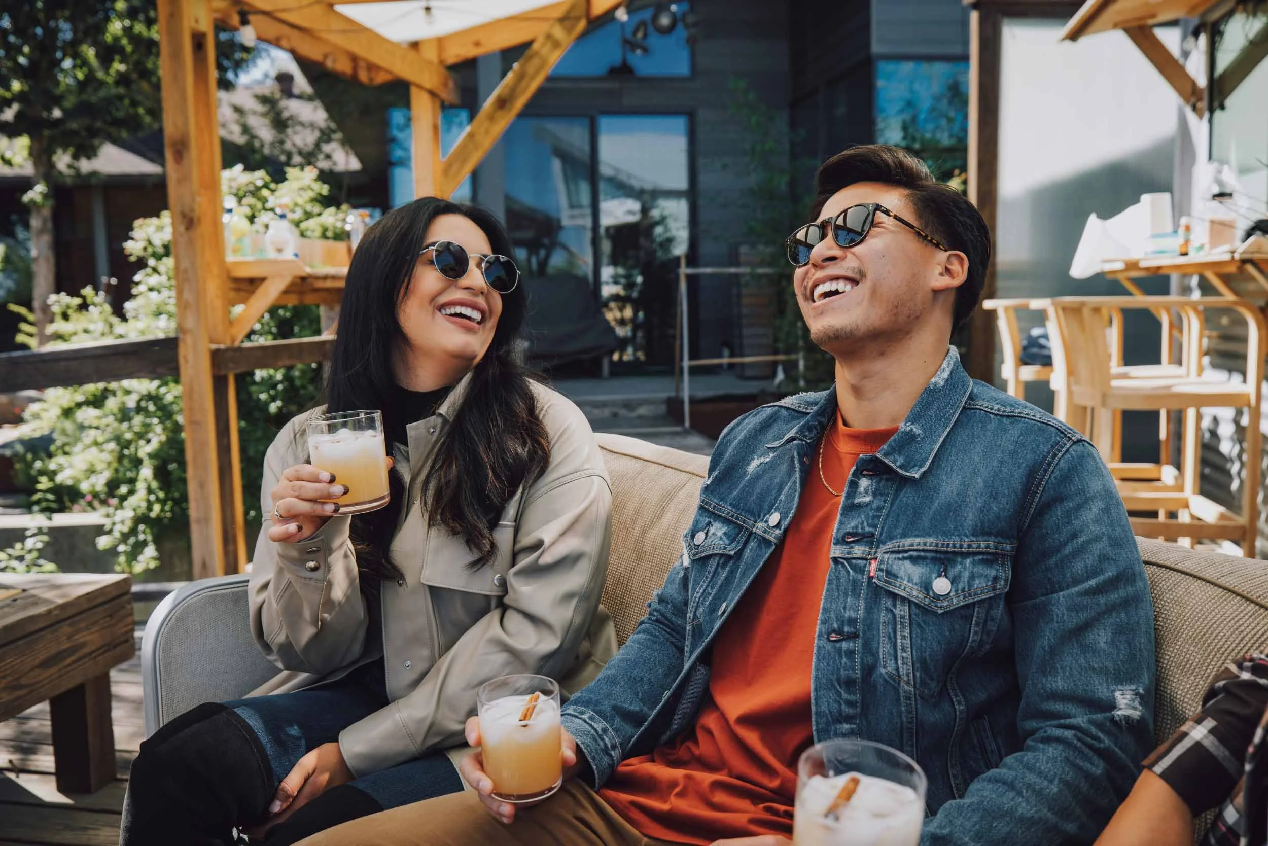 Two friends, a woman and a man, laughing and enjoying drinks on a patio during daytime. They are wearing sunglasses and casual jackets, with the woman holding a cocktail and the man holding a drink with a cinnamon stick, surrounded by outdoor furniture and greenery.
