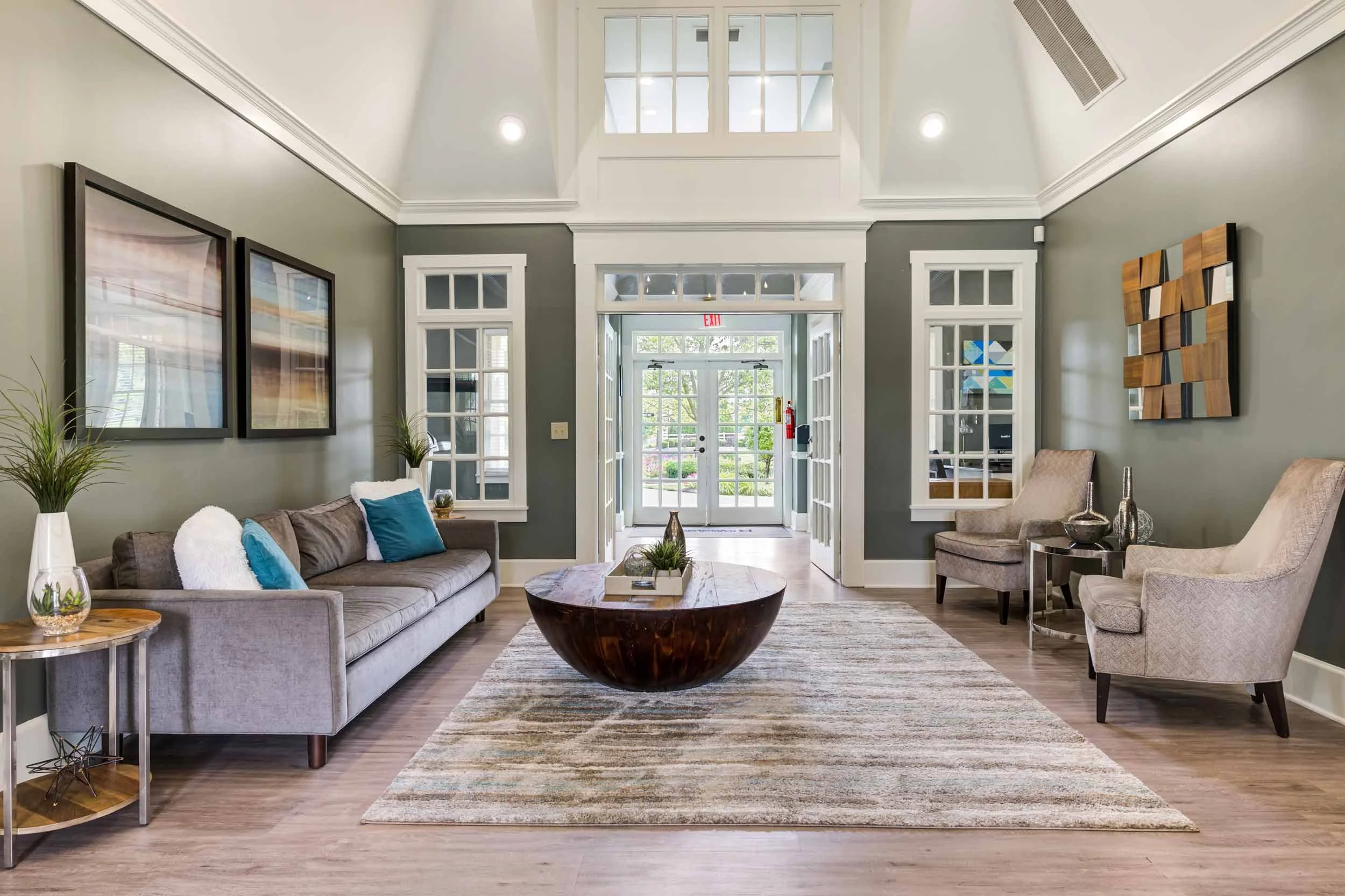 A bright, modern living room with light gray walls, white crown molding, and large windows. It features a beige sofa with blue and white pillows, two beige armchairs, a round wooden coffee table, and decorative vases. There is a textured area rug on 