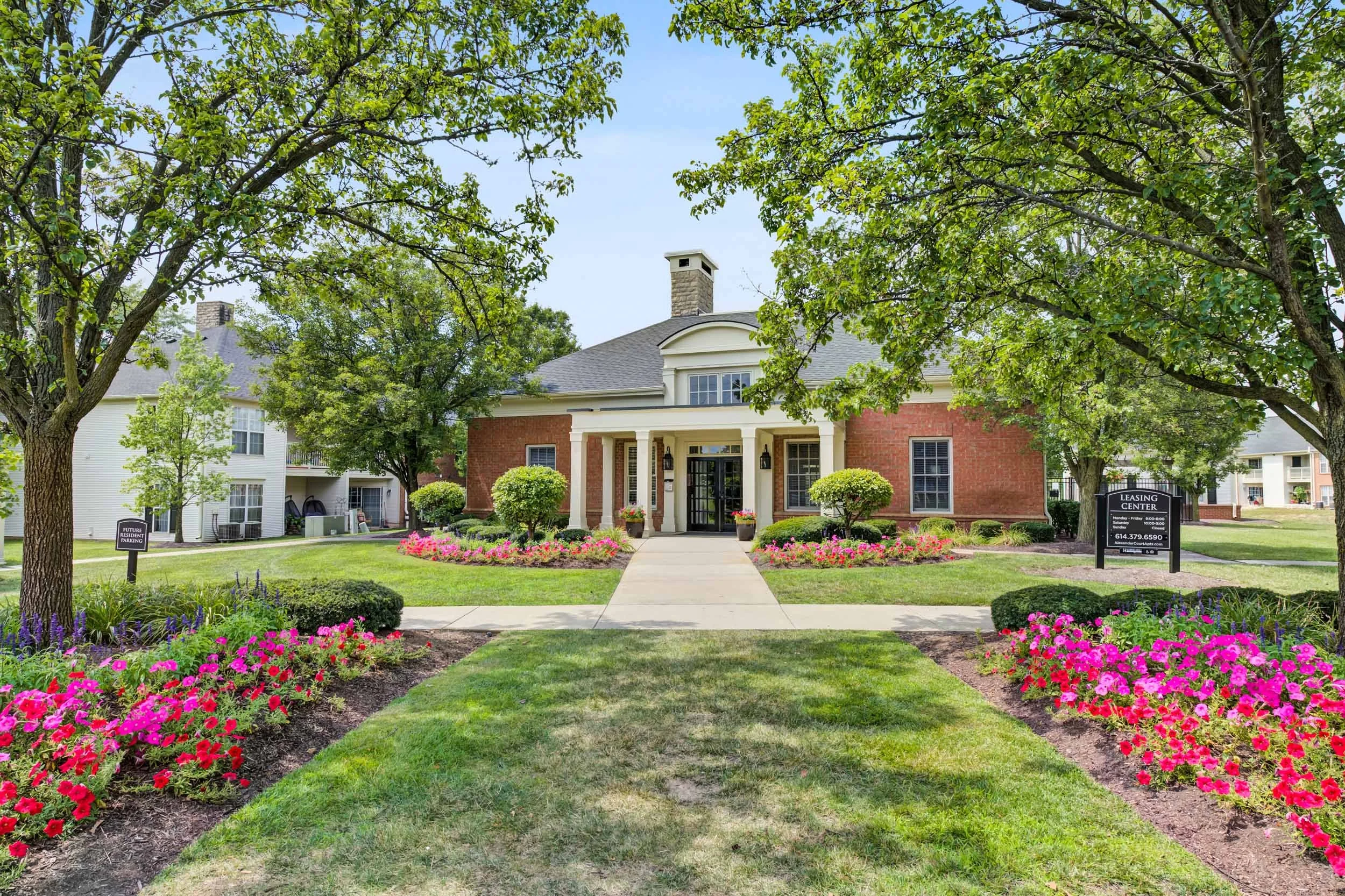 A brick residential building with a covered front porch, surrounded by a well-maintained lawn and vibrant flower beds, trees shading the area, and signs indicating leasing information.