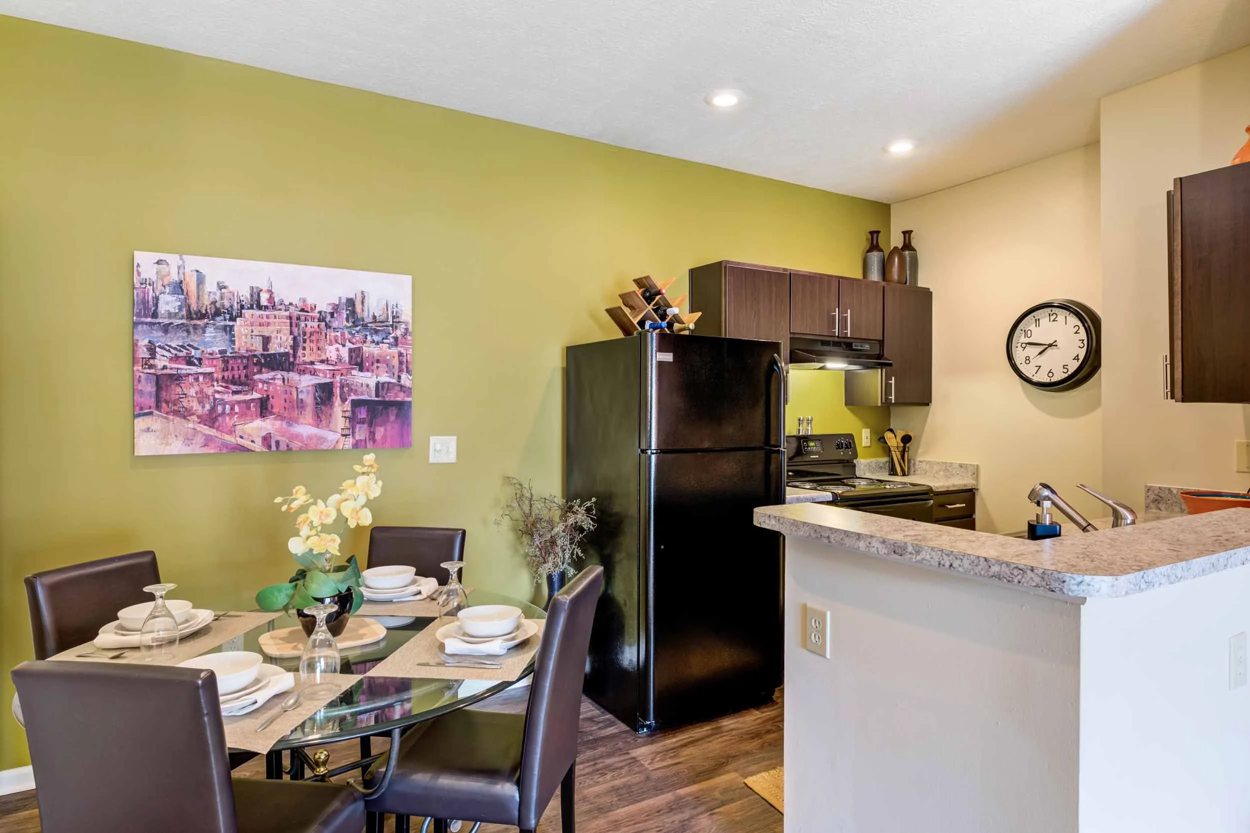 Dining area and kitchen with a green accent wall, a painting of a cityscape, a round dining table set for five, a black refrigerator, black stove, dark brown cabinets, a wall clock, and a granite countertop.