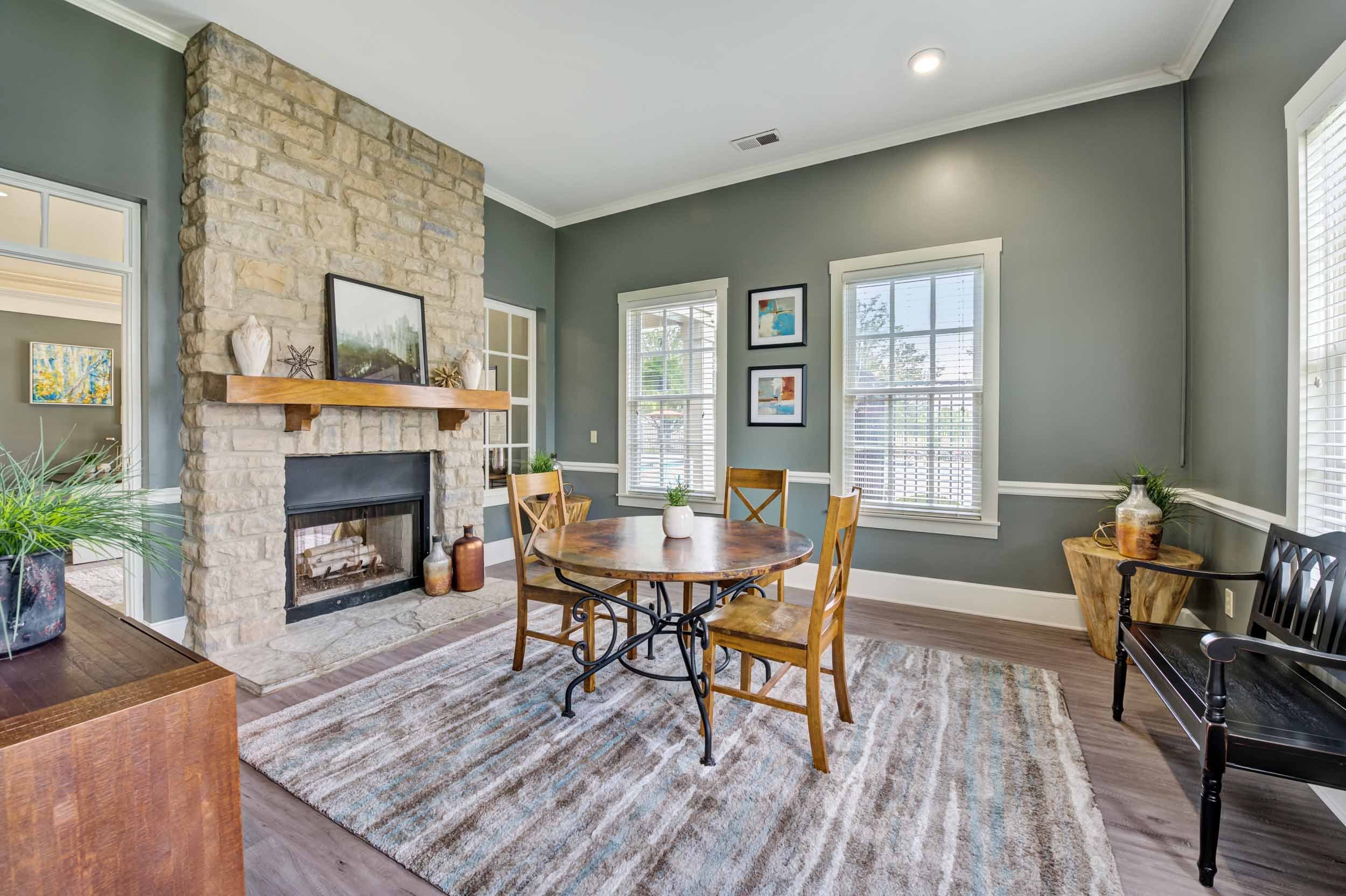 Living room with green walls, a stone fireplace with a wooden mantel, two windows with white blinds, a round wooden dining table with four chairs, a striped area rug, and decorative items like vases and framed artwork.