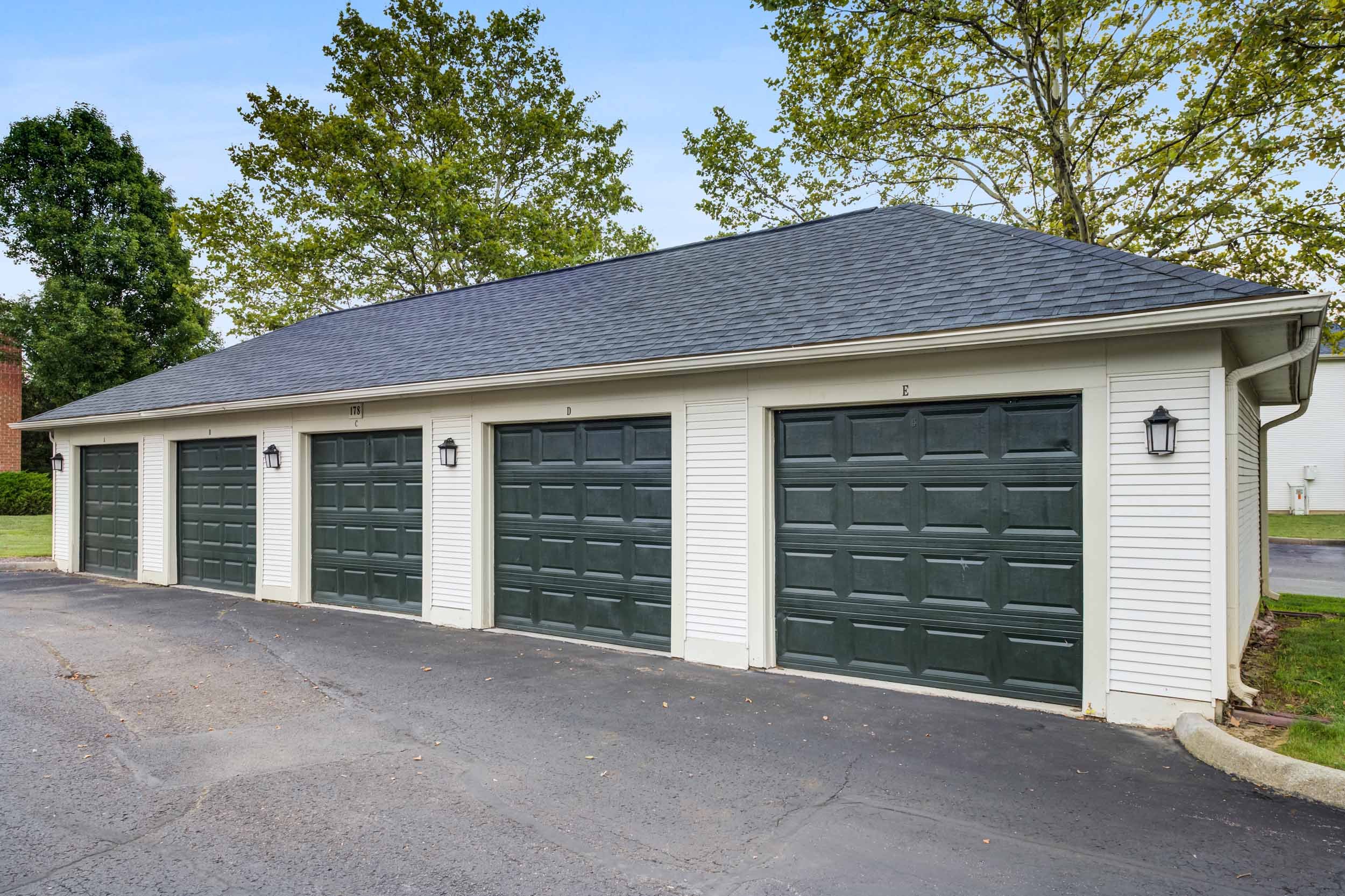 Four-car garage with white siding and black garage doors, exterior wall-mounted lights, paved driveway, green trees in the background, and a light blue sky.