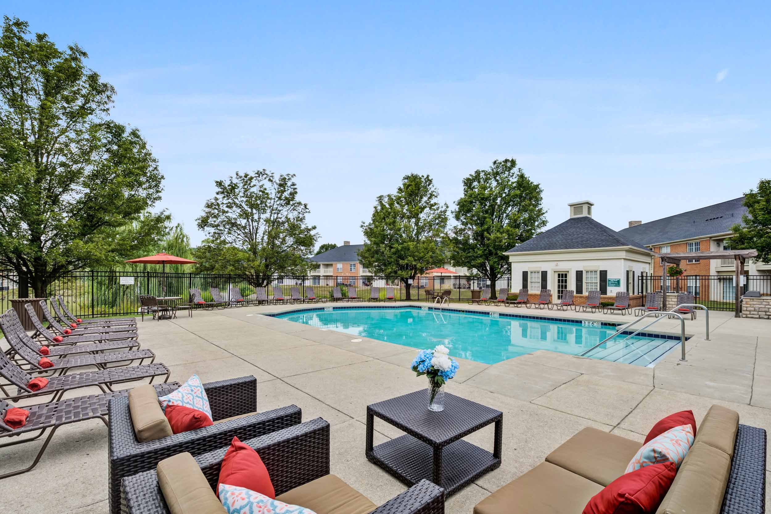 Outdoor community swimming pool with lounge chairs, umbrellas, and seating area surrounded by a black fence and trees, with residential buildings in the background.