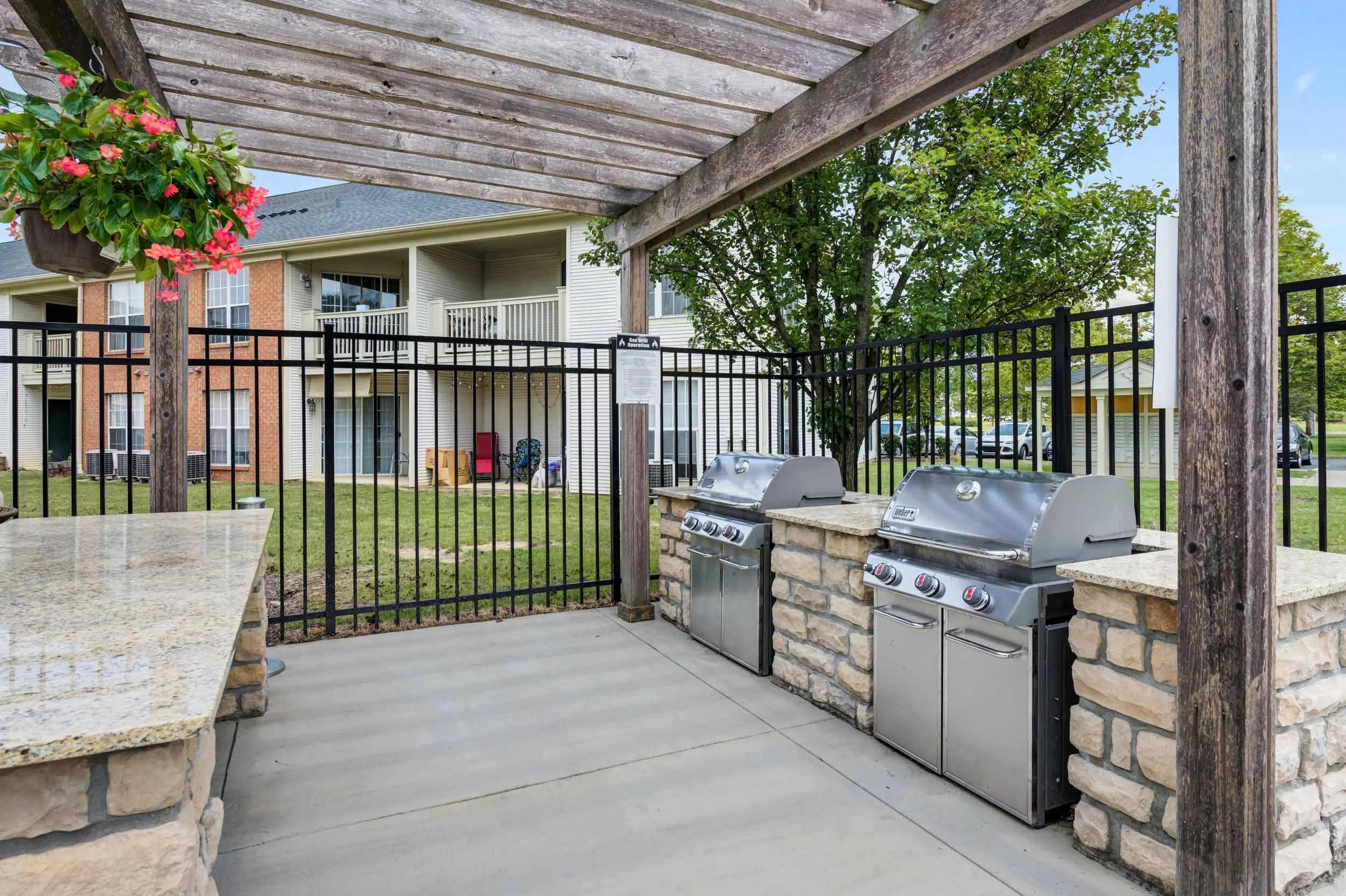 Outdoor communal area with two stainless steel grills on stone counters, surrounded by a black metal fence, beneath a wooden pergola, with an apartment building, green lawn, and trees in the background.