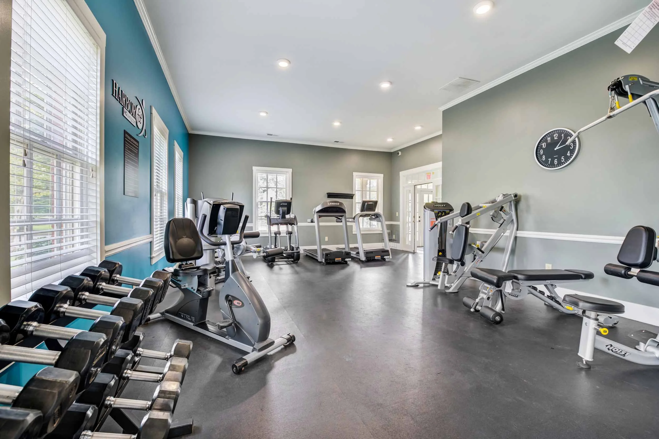 Empty gym with workout equipment including weights, exercise bikes, treadmills, and weight machines in a well-lit room with large windows