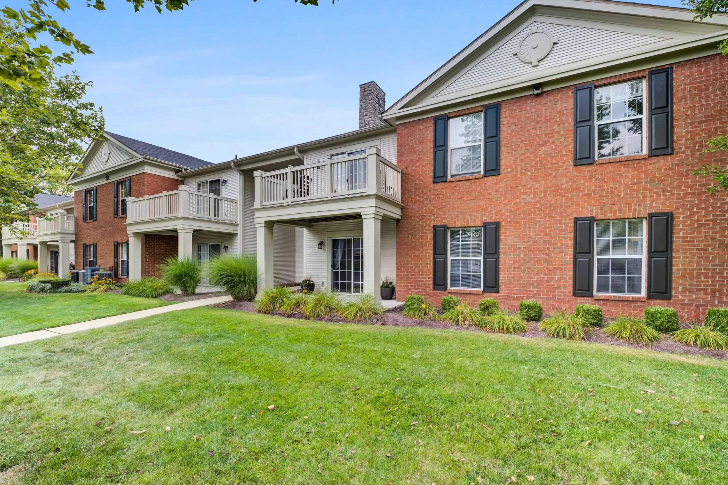 Exterior view of a multi-story apartment complex with brick and beige siding, featuring balconies with white railings, black window shutters, and landscaped grass and bushes.
