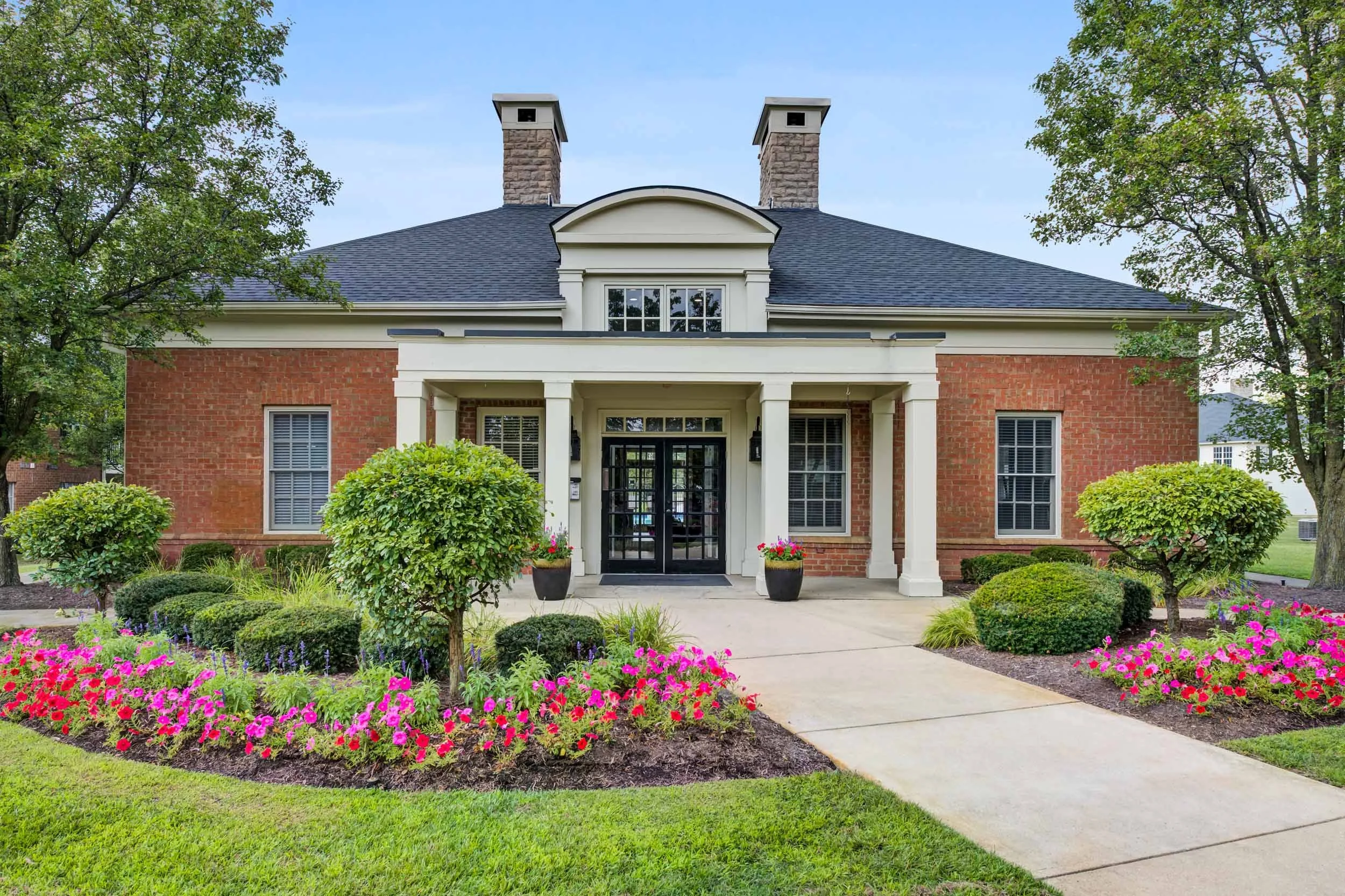 Front view of a two-story brick house with a black front door, white trim, and two chimneys, surrounded by a landscaped garden with pink flowers, green bushes, and trees.