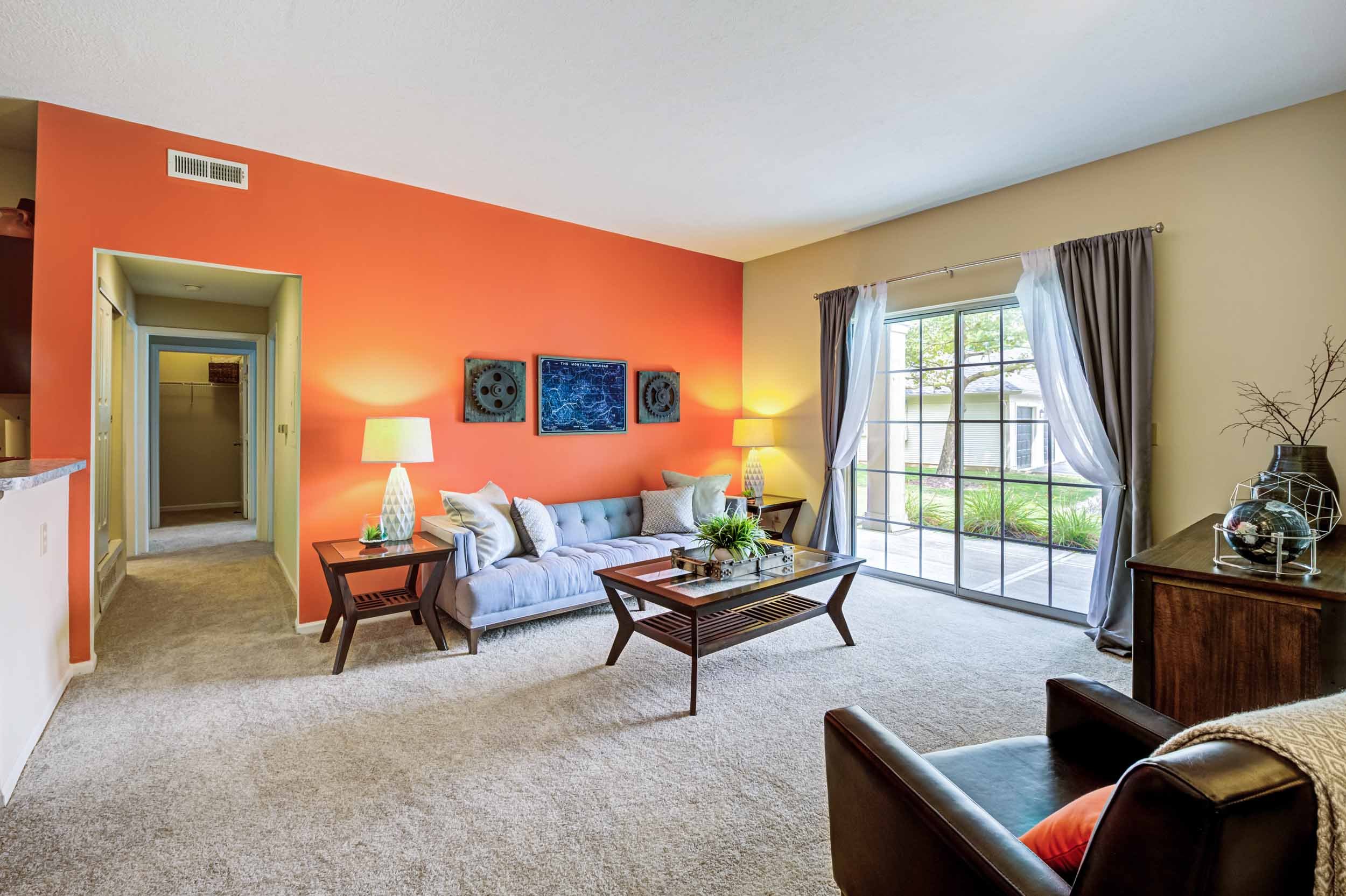 Living room with beige carpet, cream-colored wall, and an orange accent wall. Features a light blue couch with throw pillows, side tables with lamps, a wooden coffee table with a plant, and large sliding glass doors with curtains leading outside. Dec