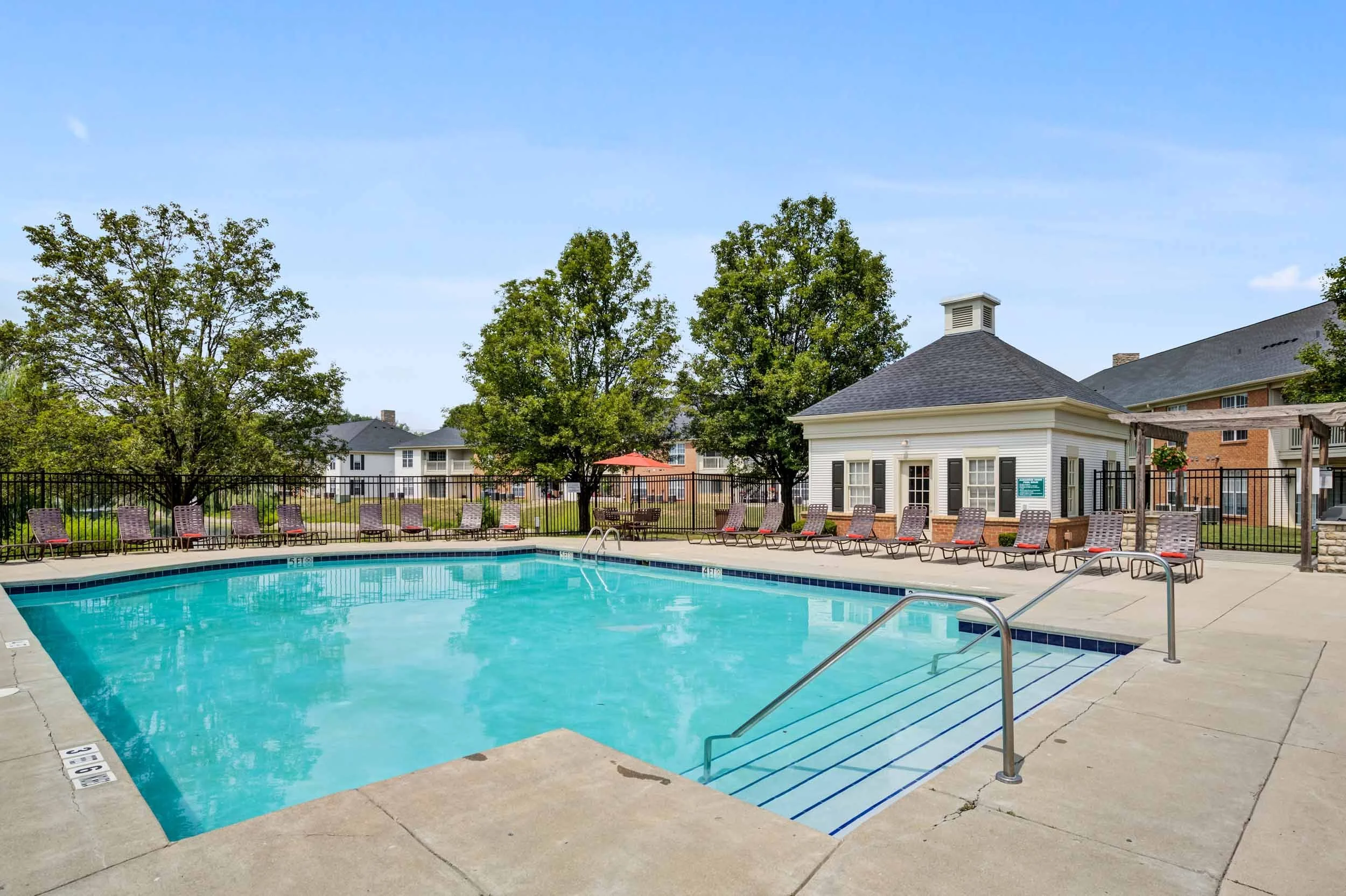 Empty outdoor swimming pool in a residential complex with lounge chairs, trees, and a white pool house under a blue sky.