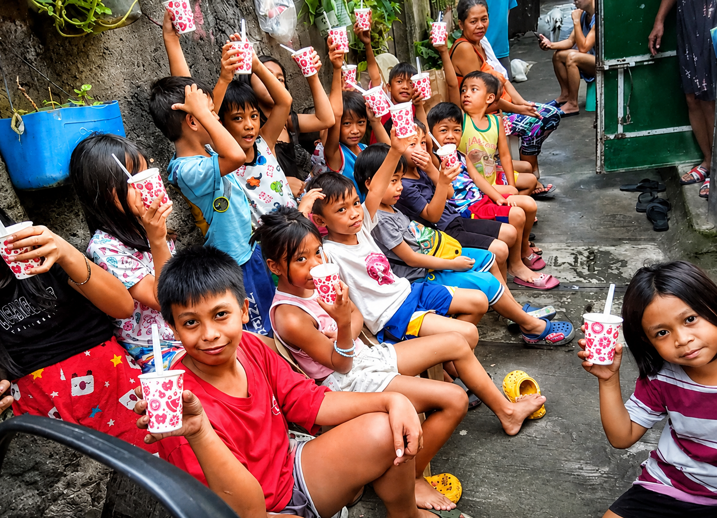 A group of children sitting on a ledge against a wall, holding colorful cups with straws, appearing to be celebrating or enjoying a party outdoors.