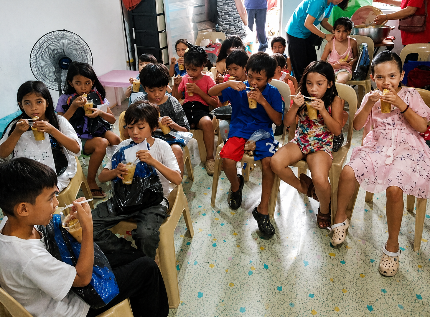 Several children sitting on plastic chairs in a room, enjoying drinks, with some adults in the background preparing food.