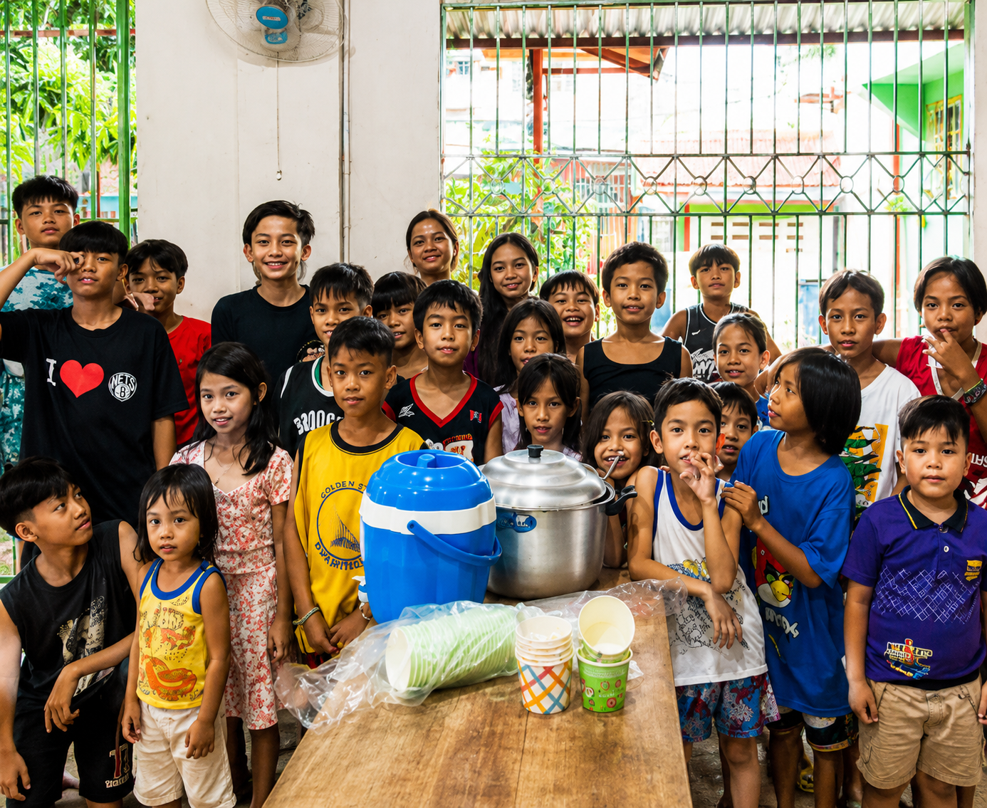 A group of children and two adults gathered behind a table with food and drinks, inside a room with open windows showing outdoor greenery.