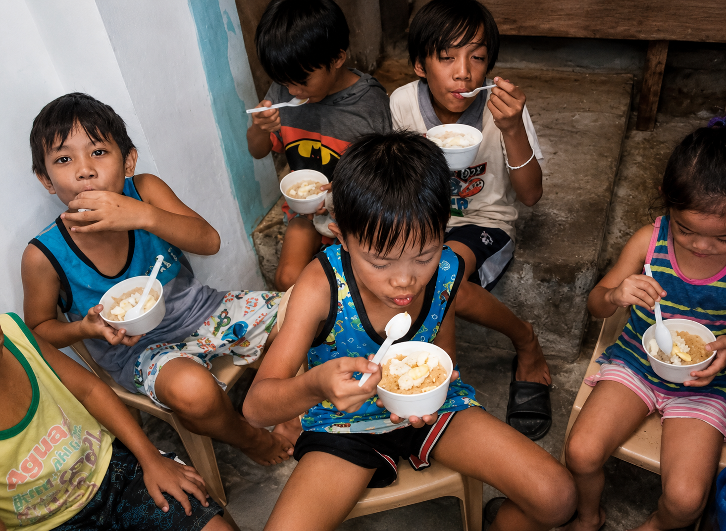 Group of children eating from bowls with spoons, sitting on small chairs in a modest room.