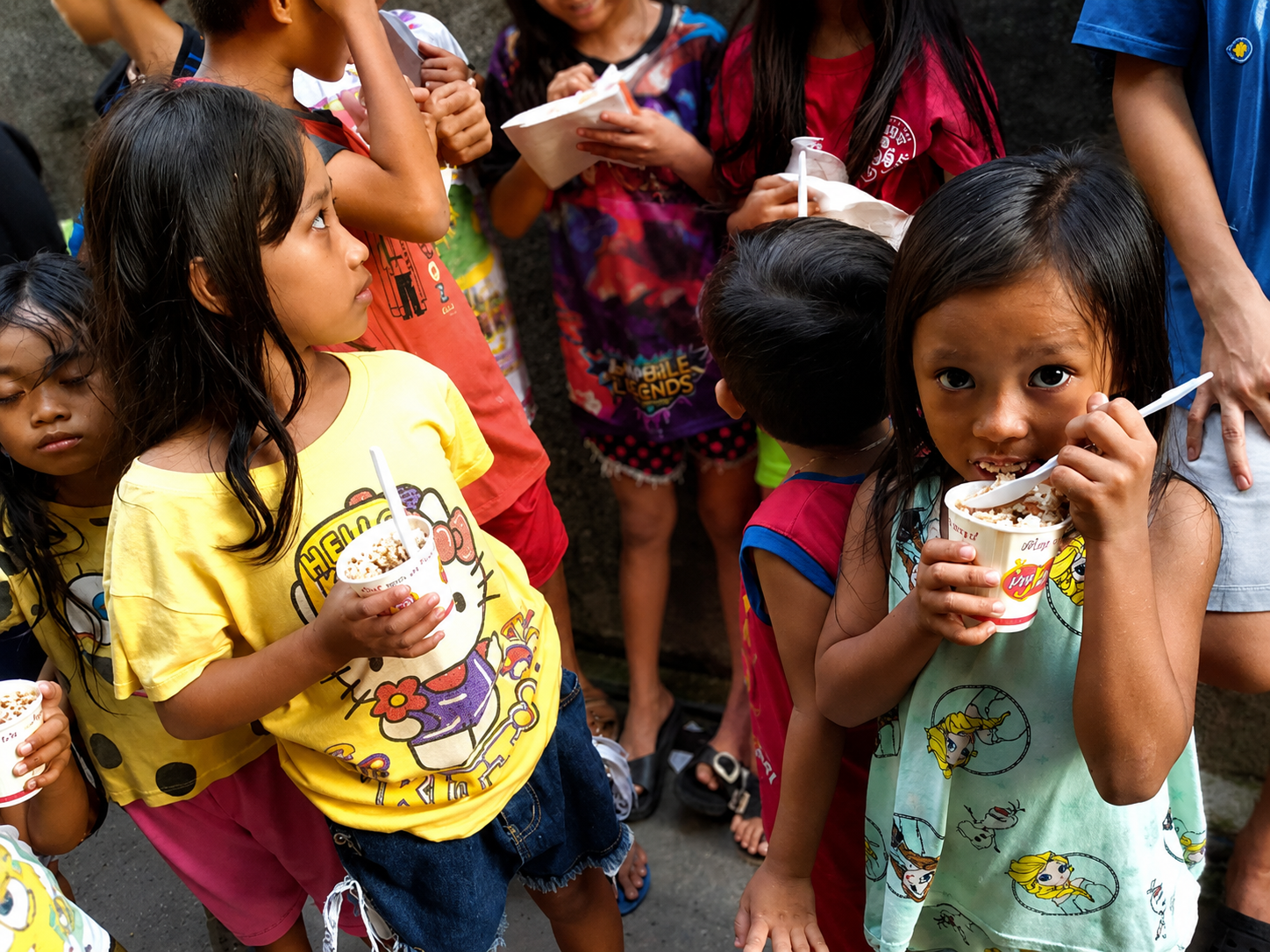 Children standing in line outdoors enjoying ice cream from cups.