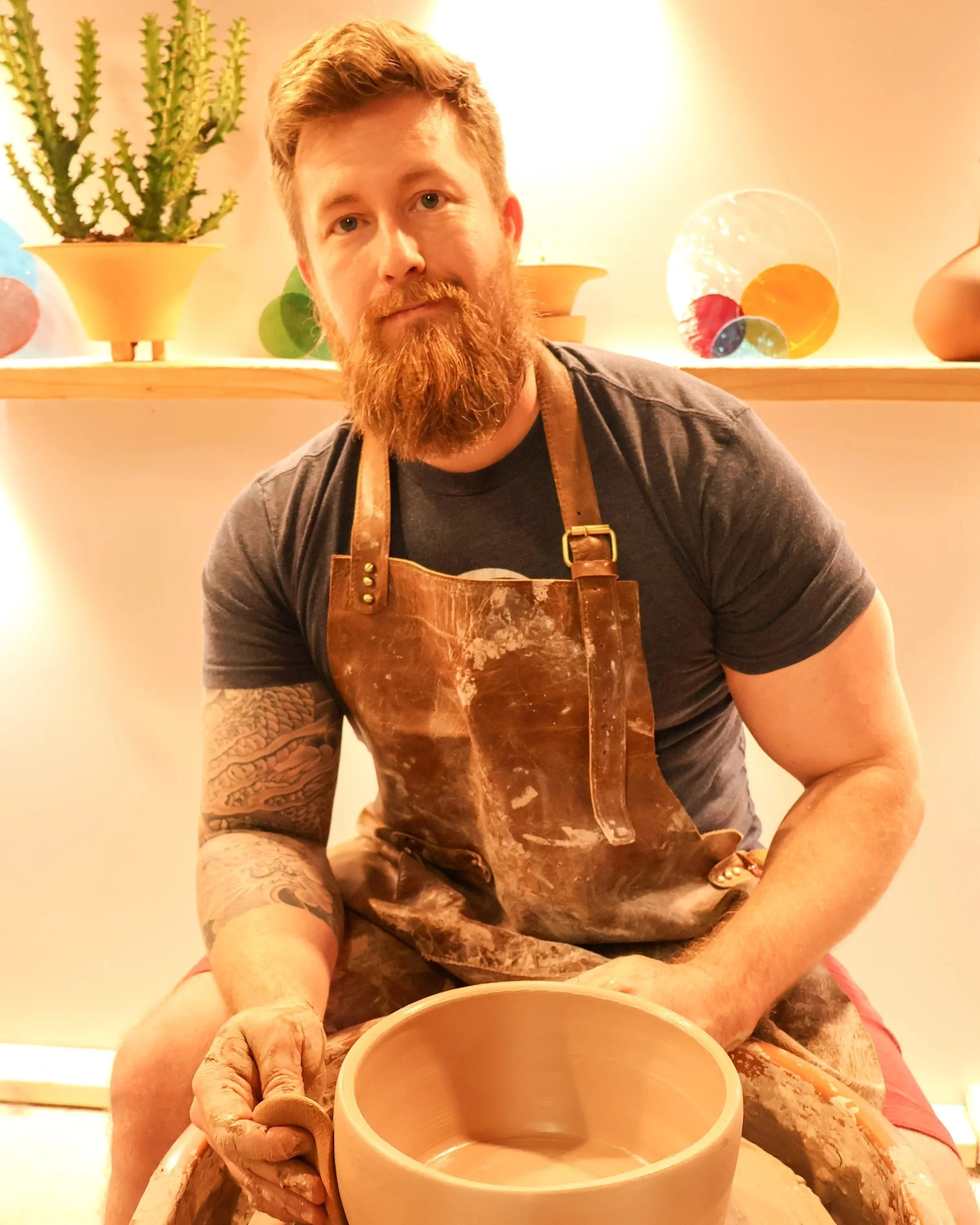Man with a beard and tattoos on his arm, wearing a leather apron, shaping clay on a pottery wheel in a ceramics studio, with decorative plants and glass art in the background.