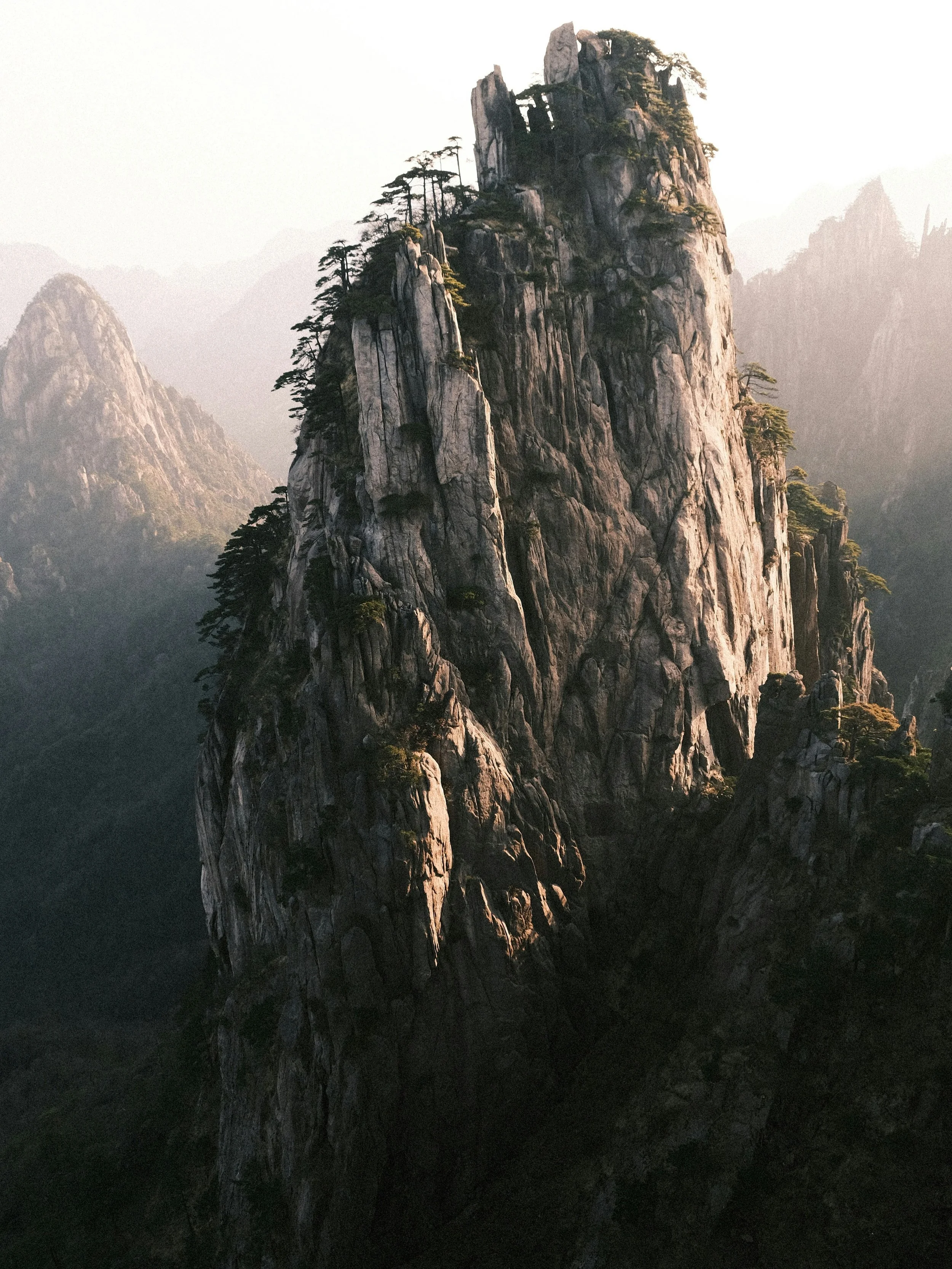 A tall, rocky mountain with sparse trees growing on its steep cliffs, partially shrouded in mist or fog. The Mountain representing the inner core strength of the Taiji practice.
