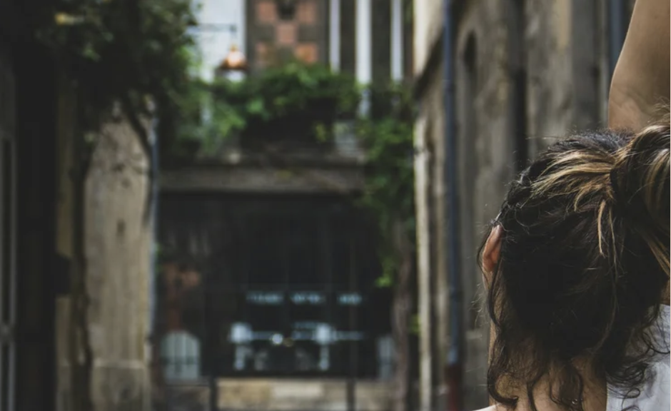Close-up of a woman with dark hair tied up, looking away in an alleyway with old buildings and greenery, evoking a feeling of what it is like at an urban yoga and meditation weekend retreat.