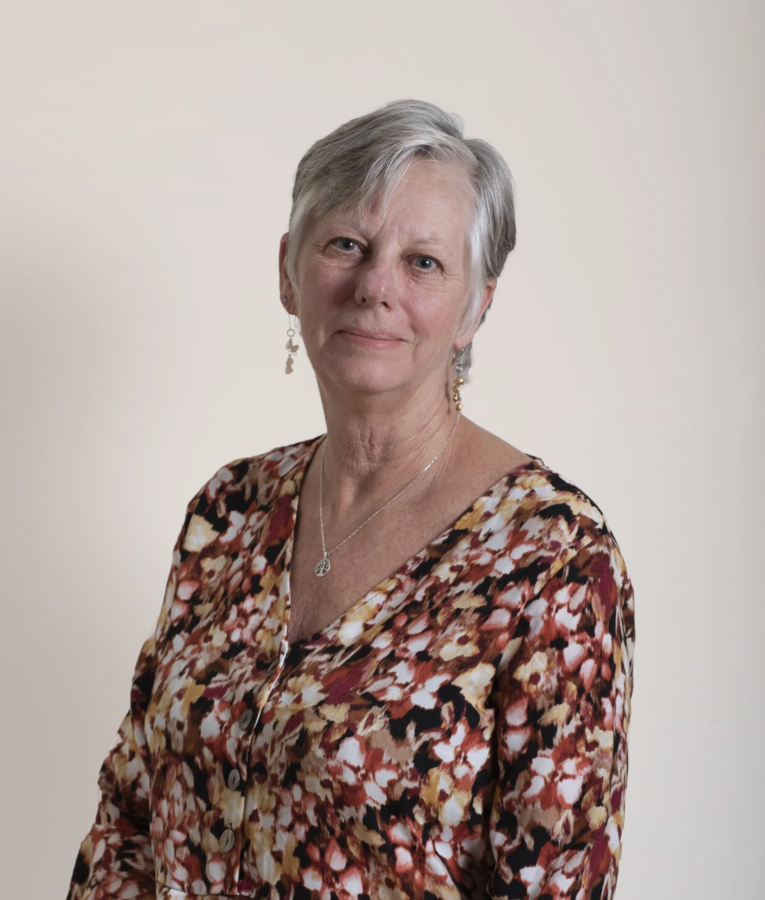 A portrait of a woman with short gray hair, wearing a floral-patterned blouse, earrings, and a necklace, standing against a plain light background.