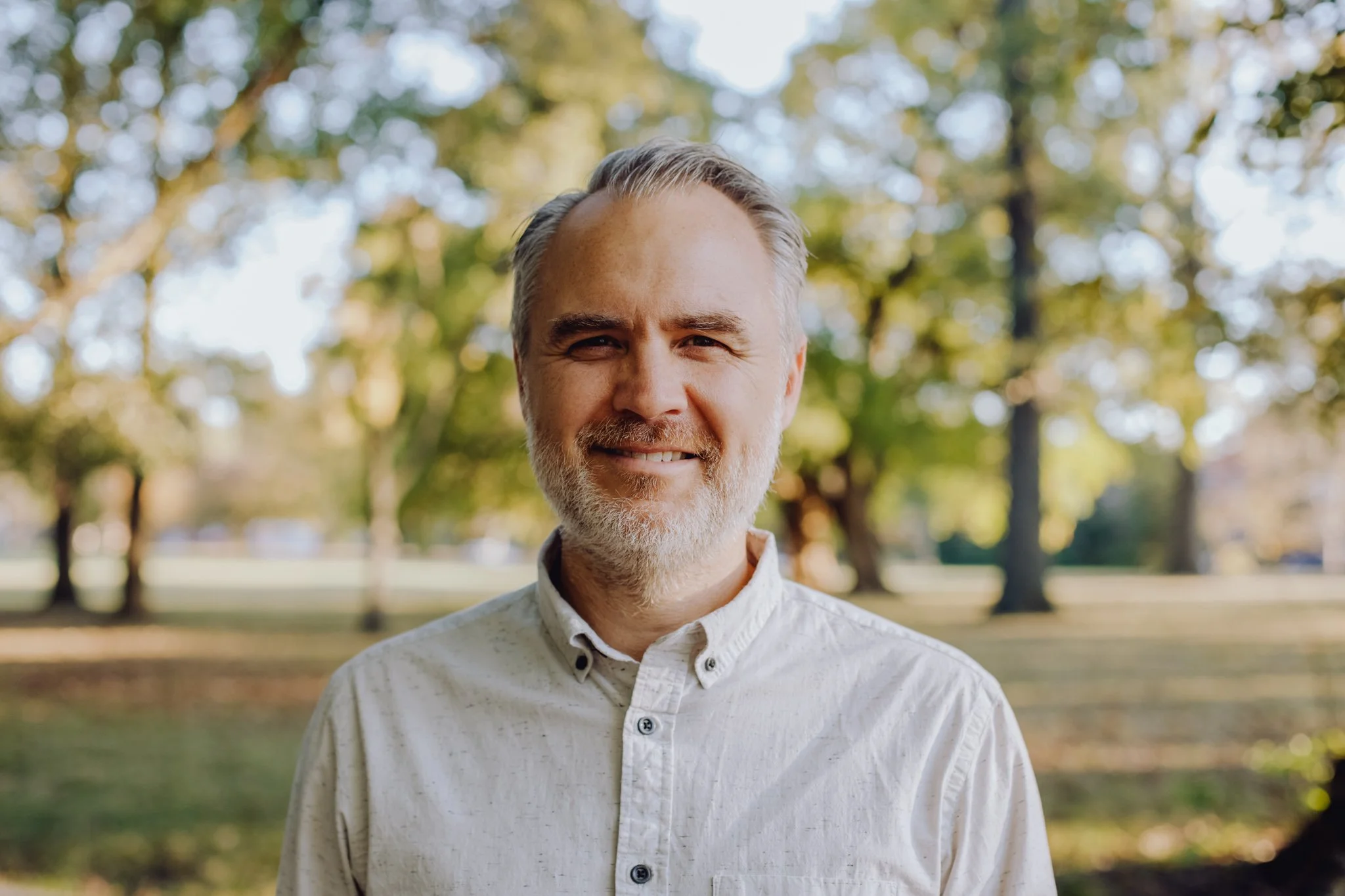 A smiling man with gray hair and beard wearing a light-colored shirt, standing outdoors with trees and sunlight in the background.