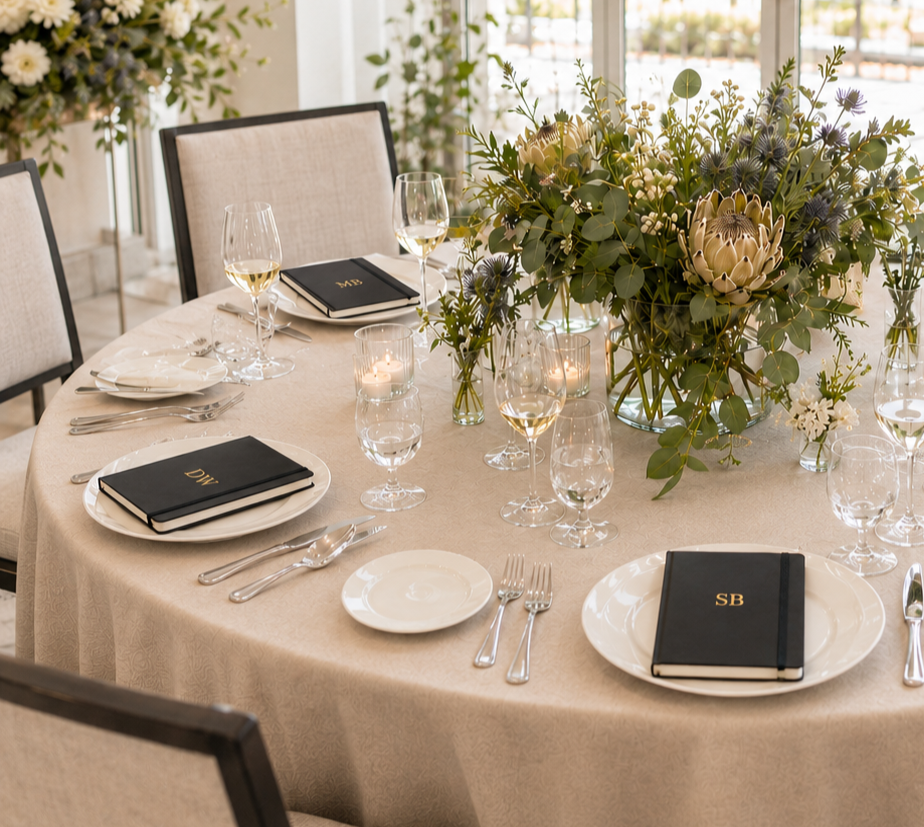 Elegant dining table set with white plates, silverware, wine glasses, and black monogrammed napkins, centered around a large floral arrangement with white and purple flowers and greenery.