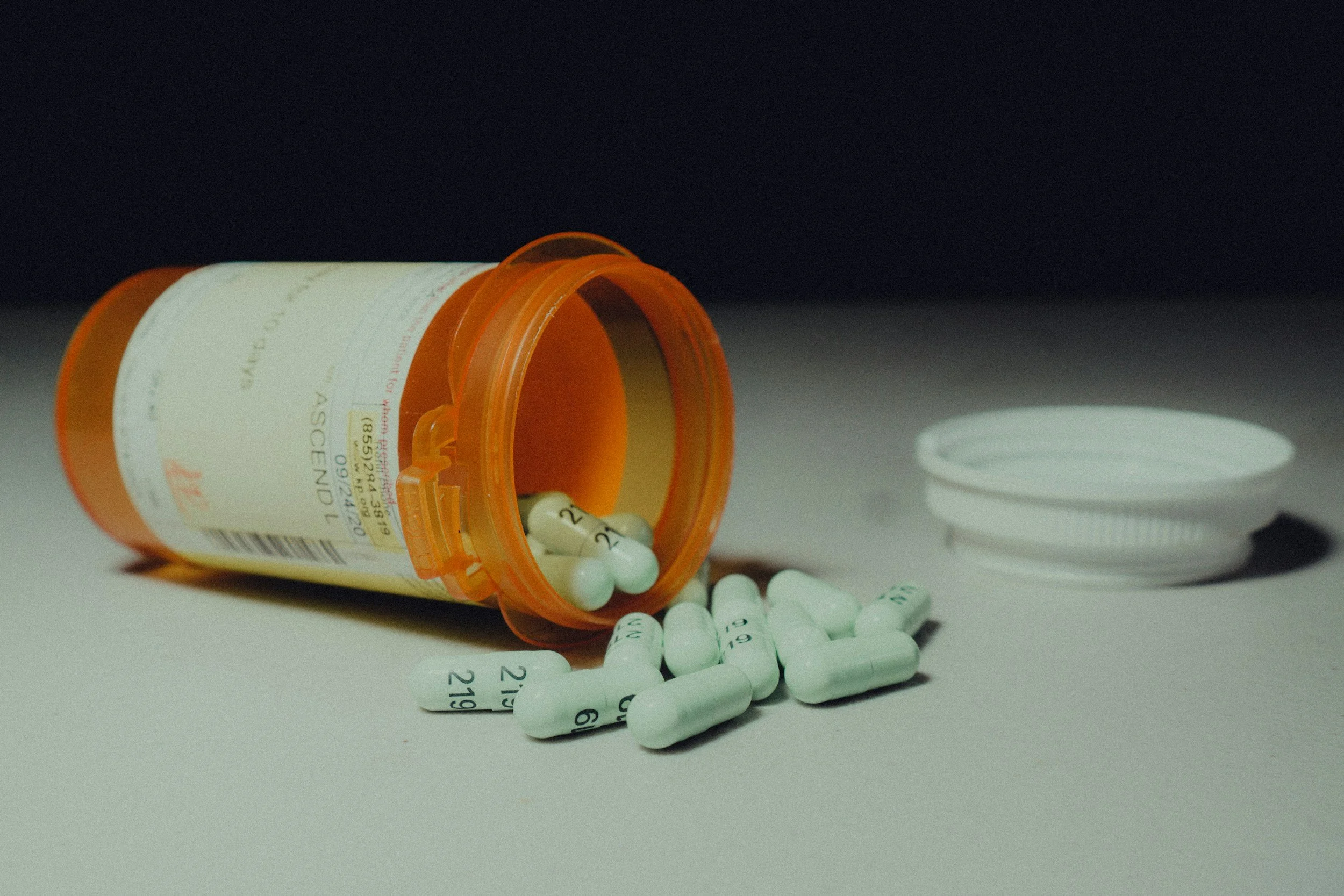 An orange prescription pill bottle is tilted on its side with white tablets spilling out, some tablets have black numbers printed on them, and the bottle has a white label with red and black text.