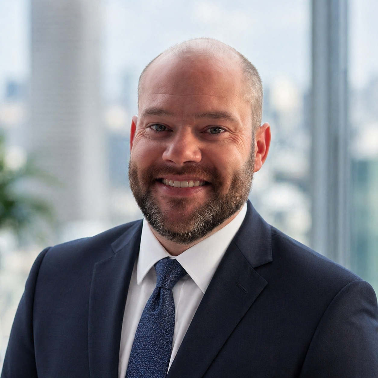 A man with a beard and bald head wearing a dark suit, white shirt, and blue tie, smiling in an office with large windows and a cityscape background.